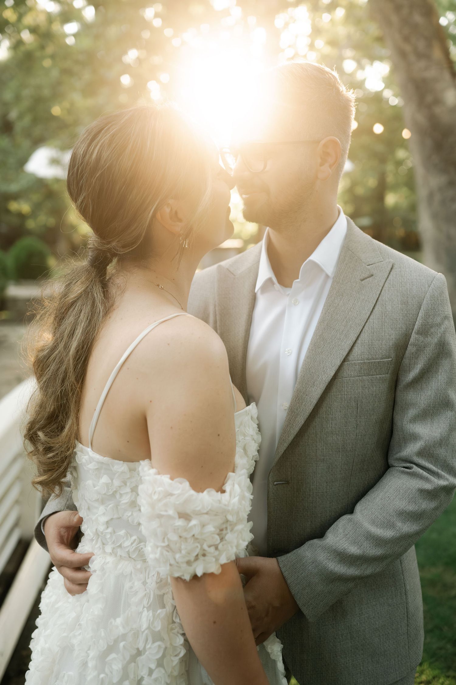 A romantic wedding couple embraces in soft sunlight filtering through trees during their outdoor photo session.