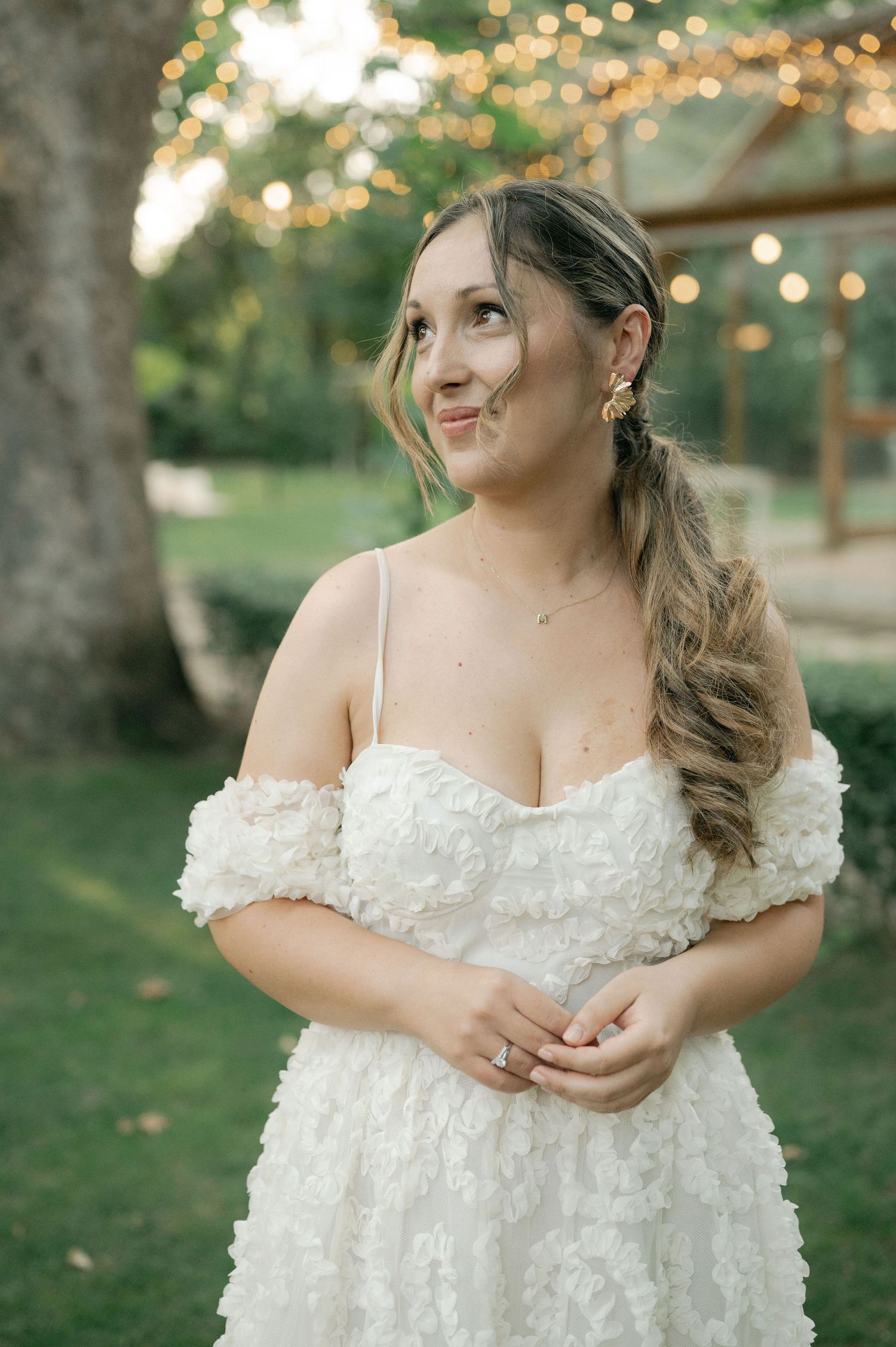 A bride in a delicate white gown stands in a garden setting with twinkling lights and natural greenery behind her.