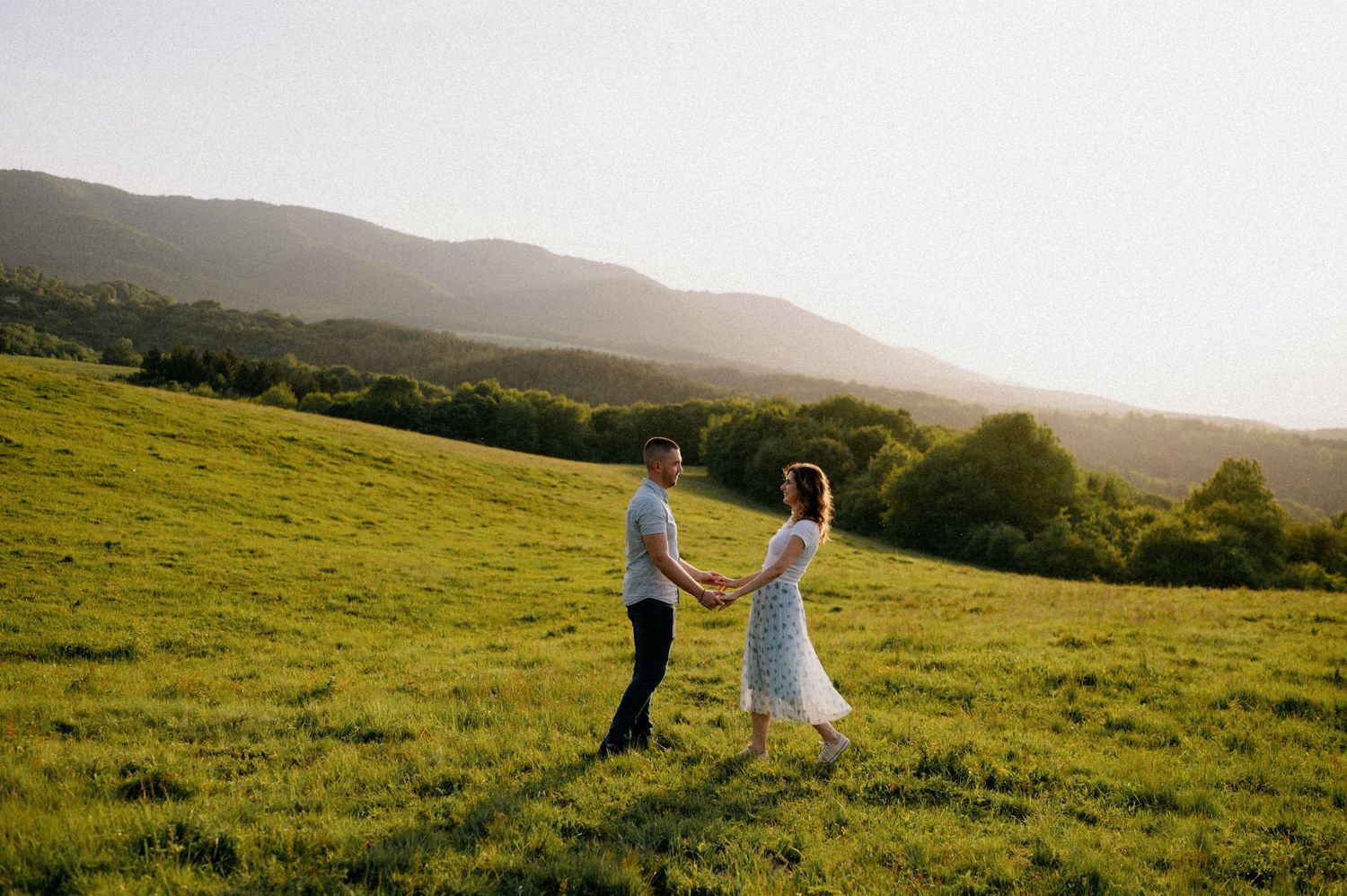 Two people hold hands while walking through a scenic hillside meadow at sunset.