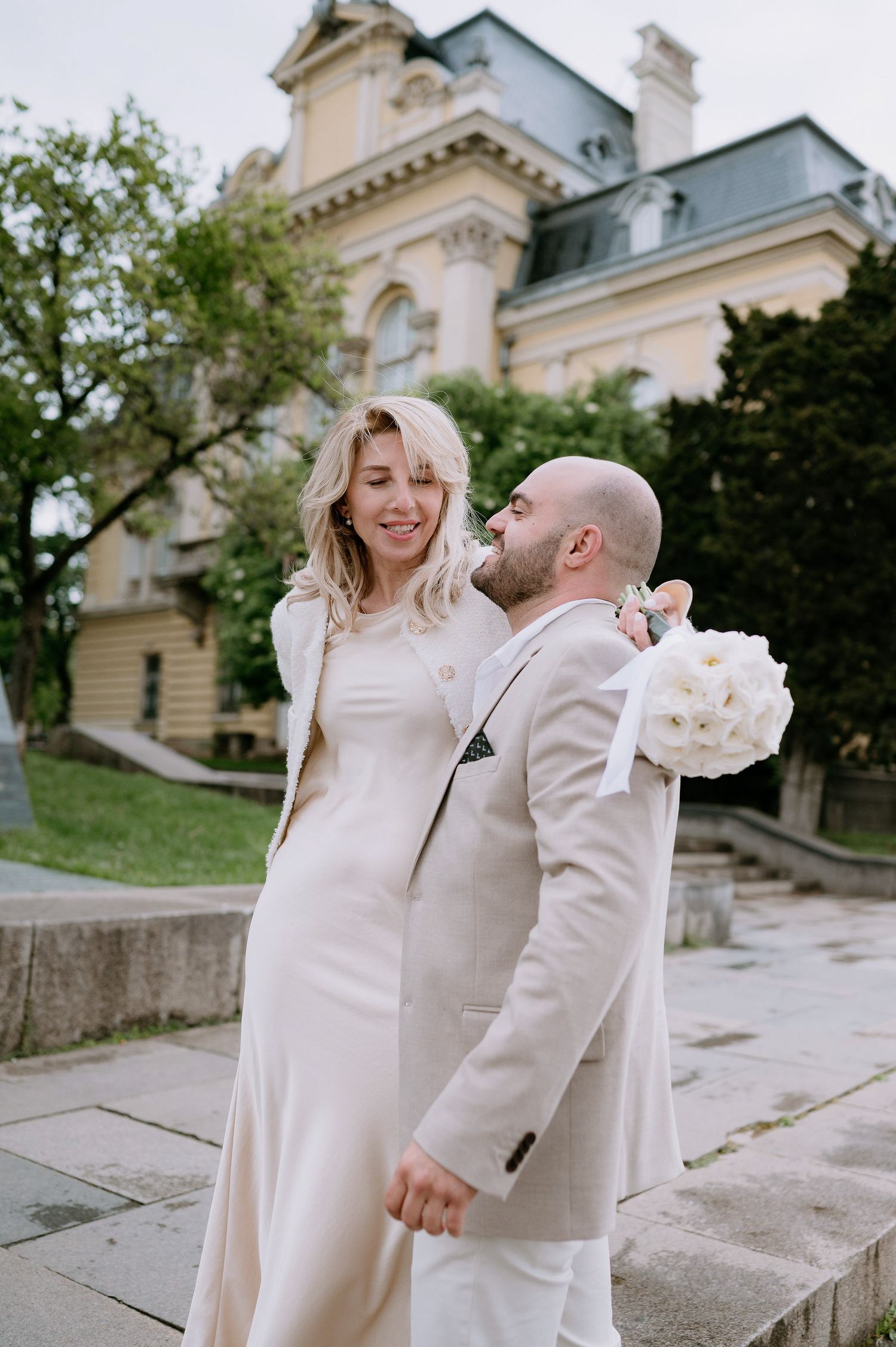 A couple wearing matching light-colored outfits embrace and laugh together on stone steps.