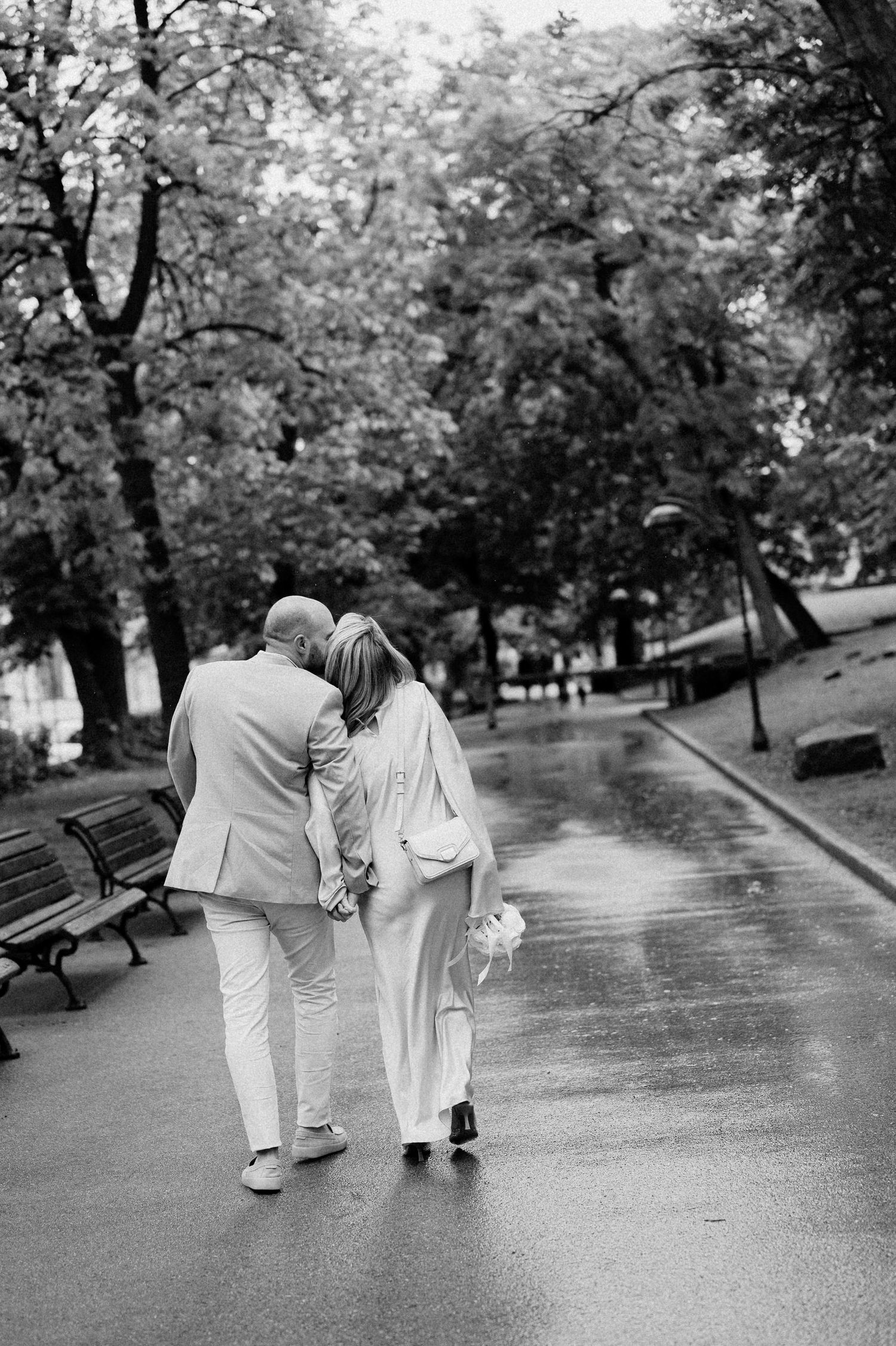 A couple in matching light-colored outfits walks together along a tree-lined path in a park.
