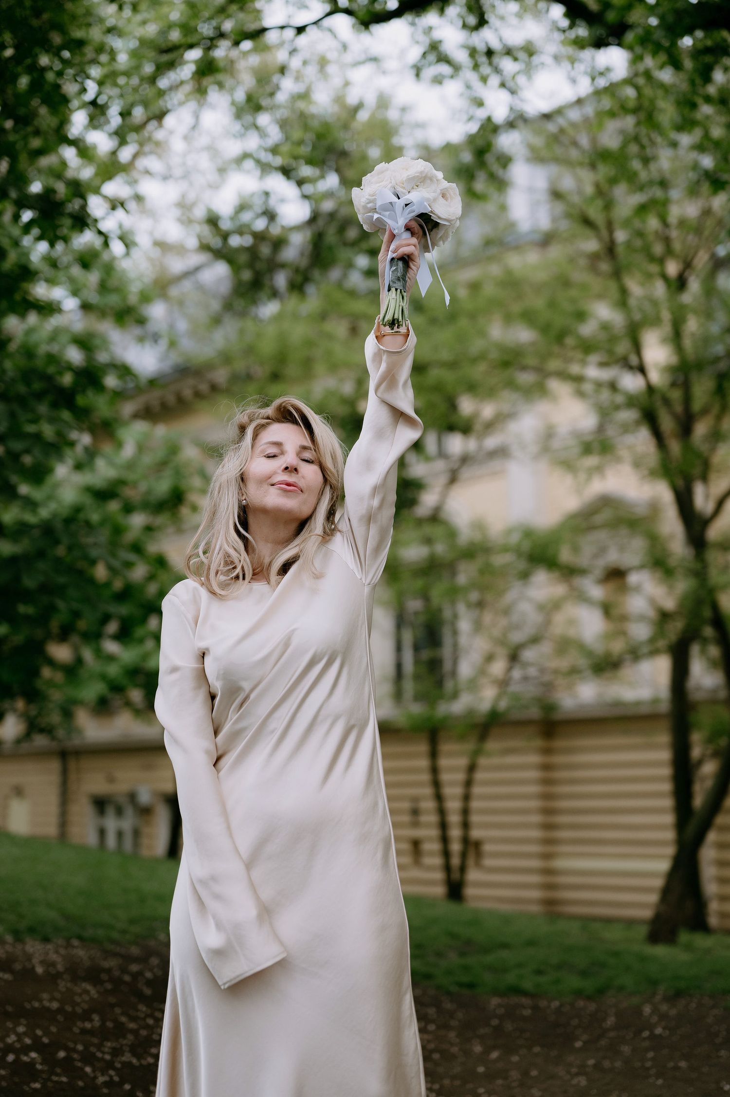 A figure in a cream-colored dress raises a bouquet against a green garden backdrop.
