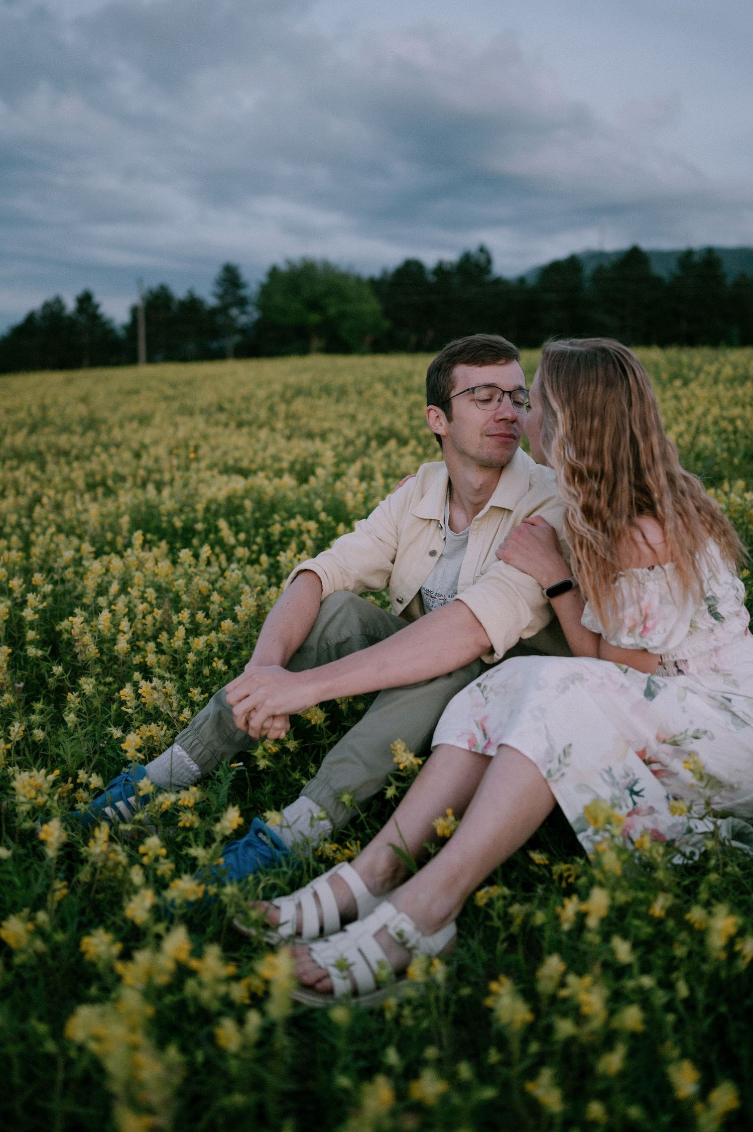 A romantic couple lounges together in a blooming yellow flower field on an overcast day.