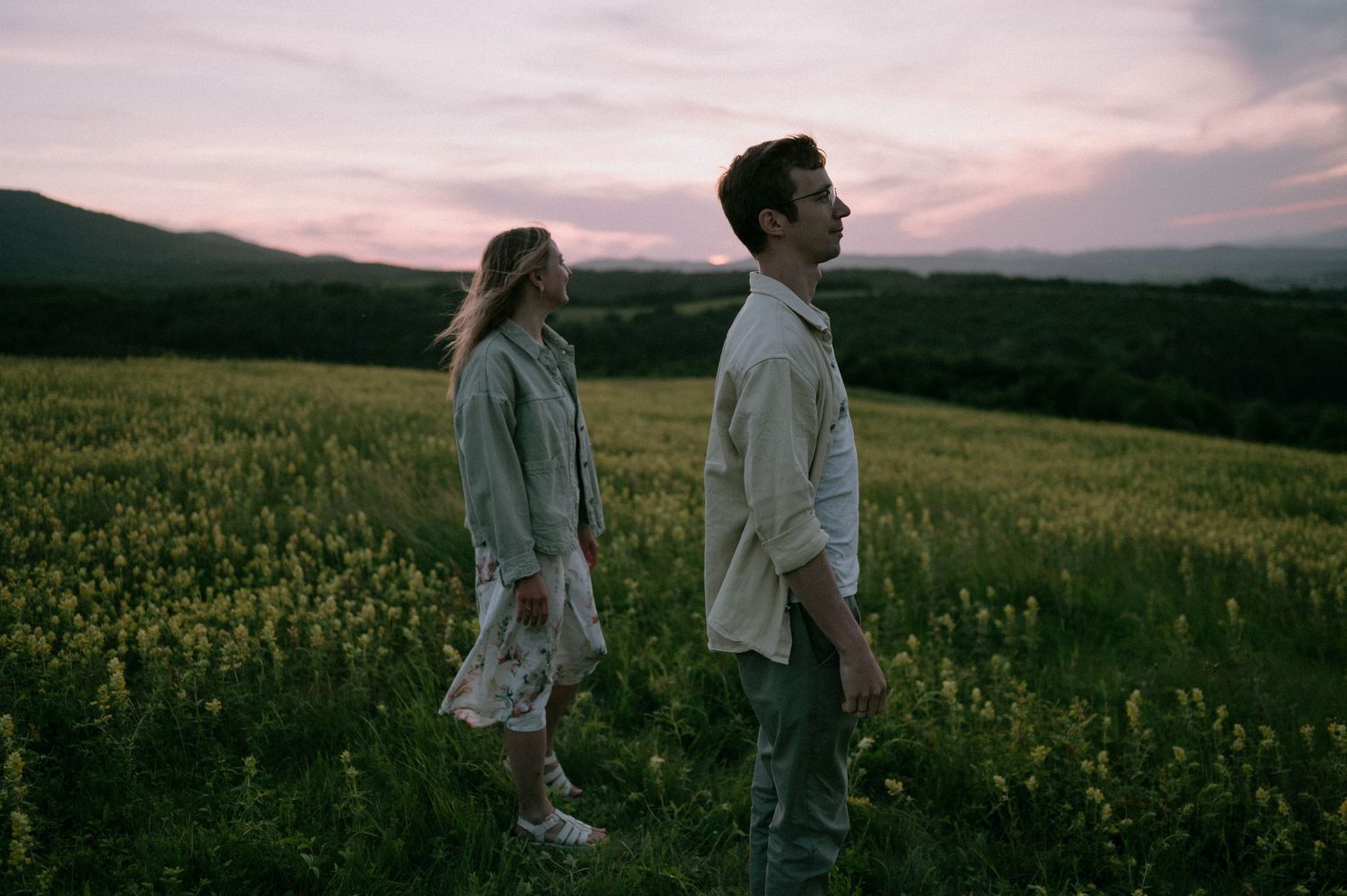 Two people stroll through a scenic meadow with mountains and pink sky in the background.