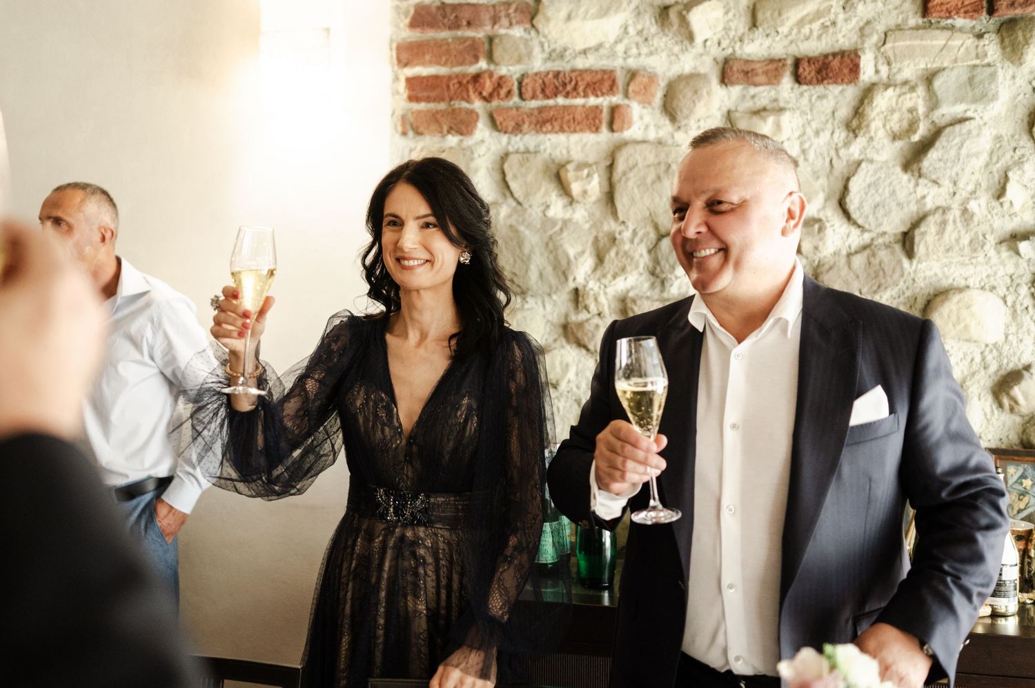 Wedding guests share laughs and drinks against a rustic stone wall backdrop.