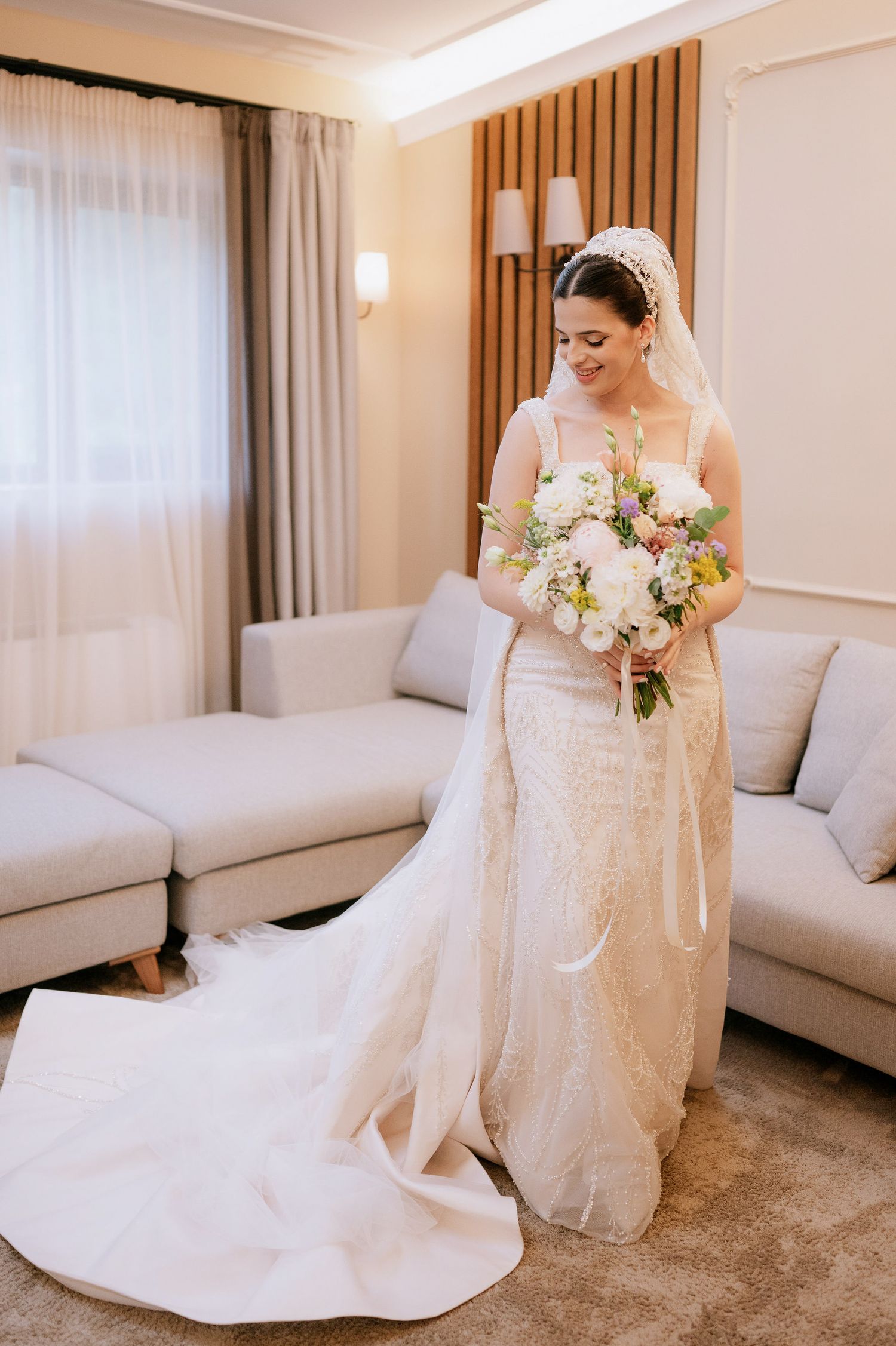 Bride in classic white gown holds a soft pastel bouquet while standing in a modern room with wood paneling.