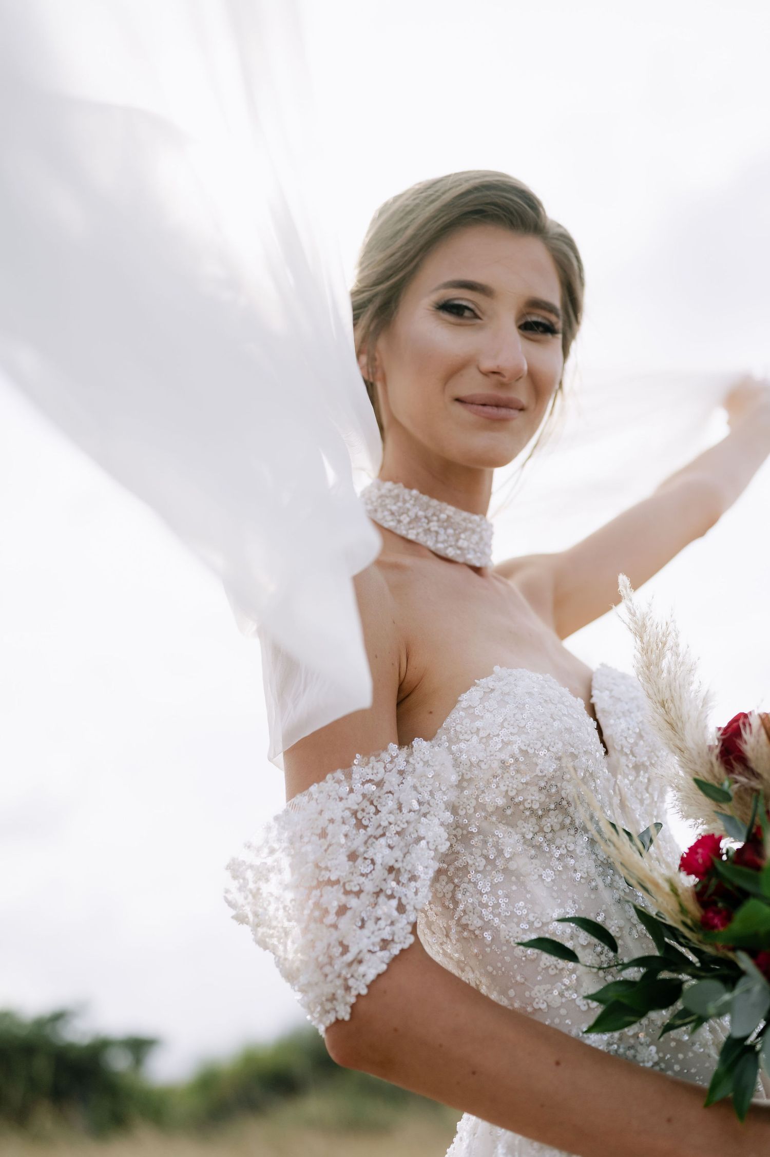 A bride in an off-shoulder wedding dress poses with her veil and holds a romantic mixed flower bouquet.