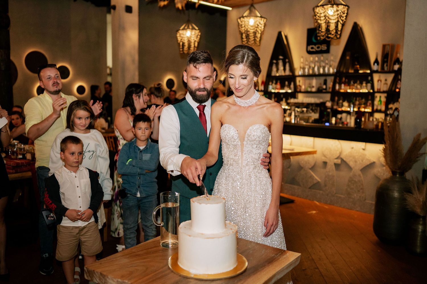 Wedding celebration in a warmly lit venue with guests surrounding the cake table.