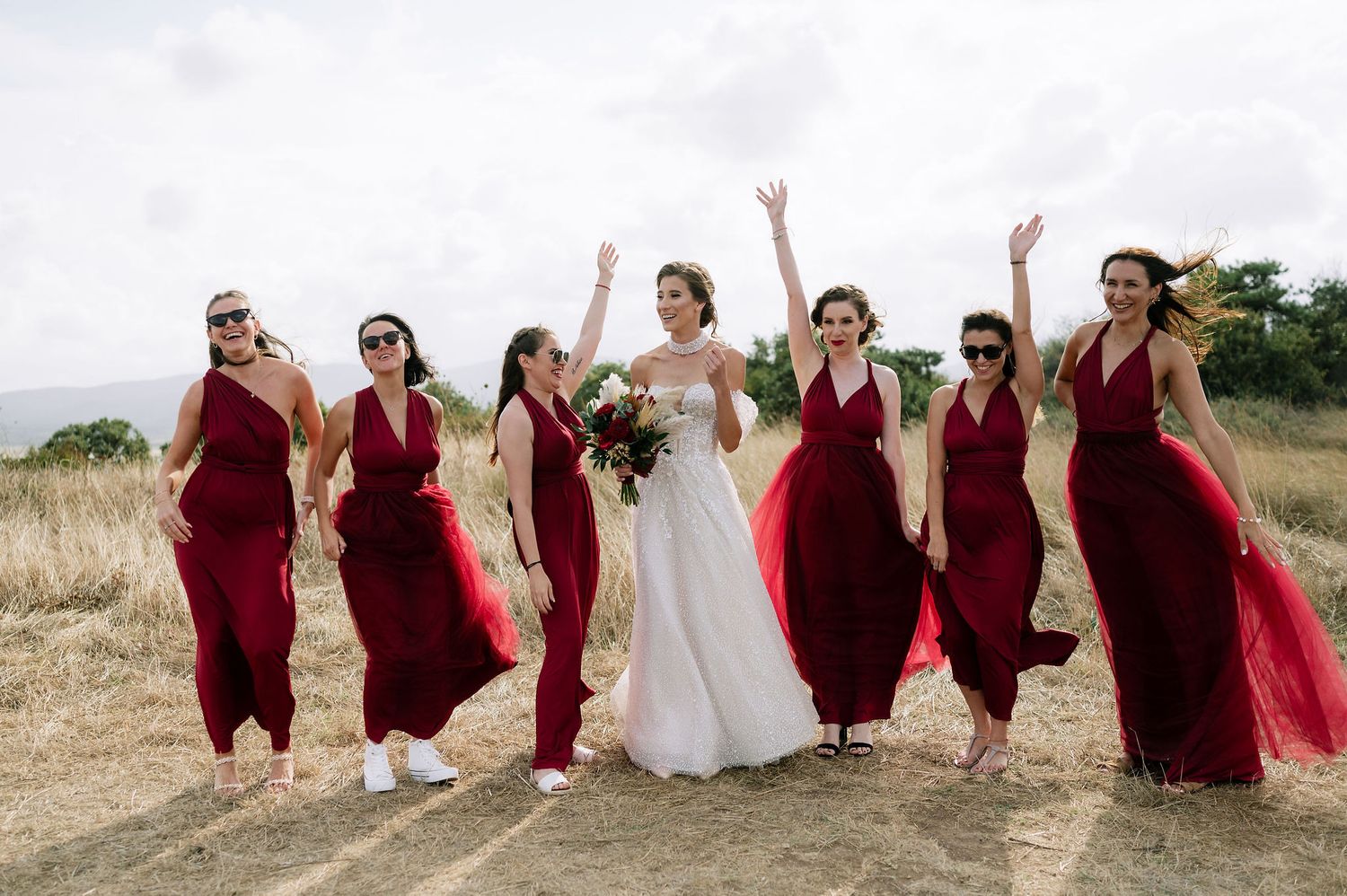 Wedding party in burgundy dresses poses playfully on a grassy hill.