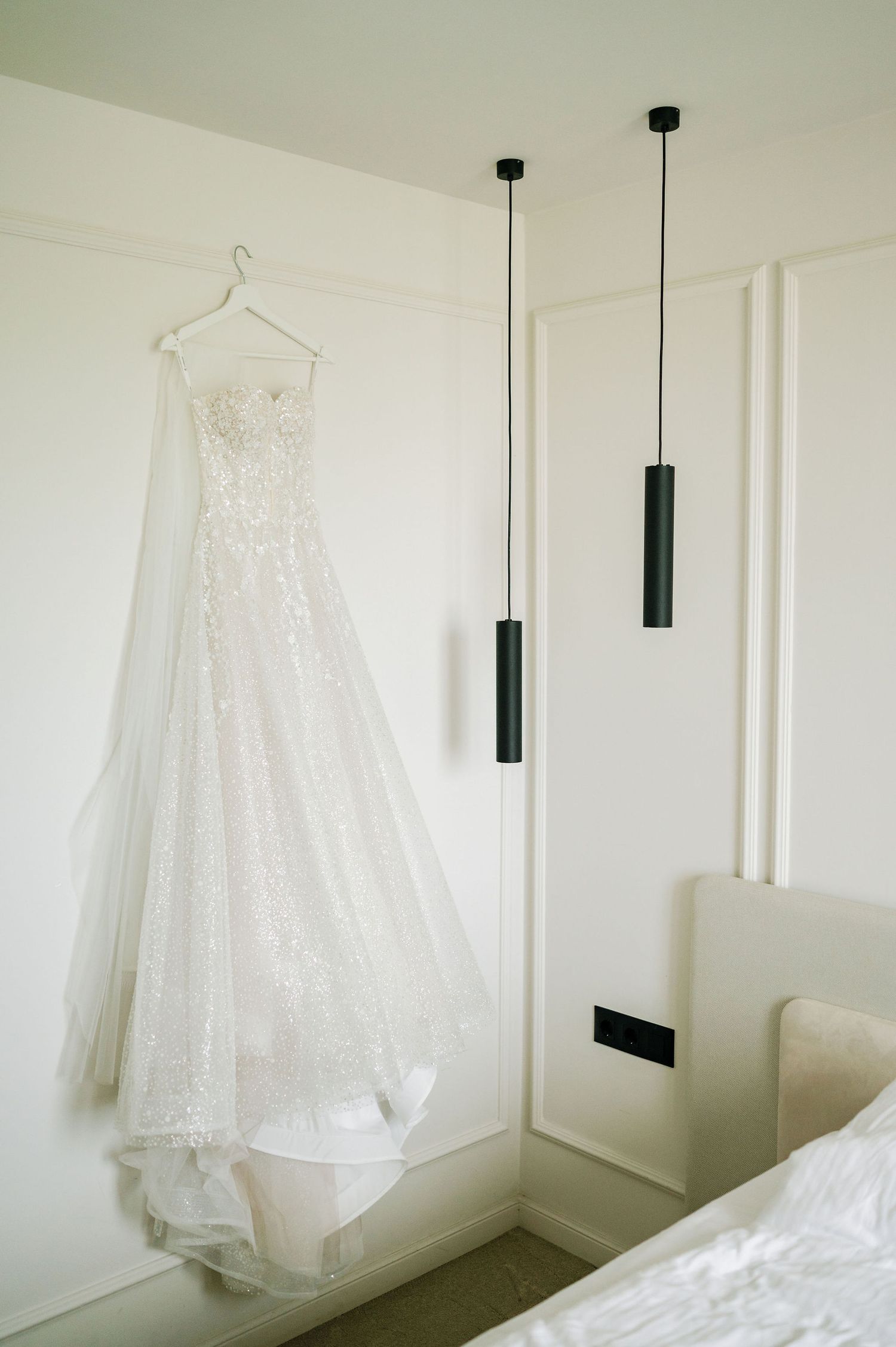 White lace wedding dress hanging elegantly against minimalist white wall with pendant lights.