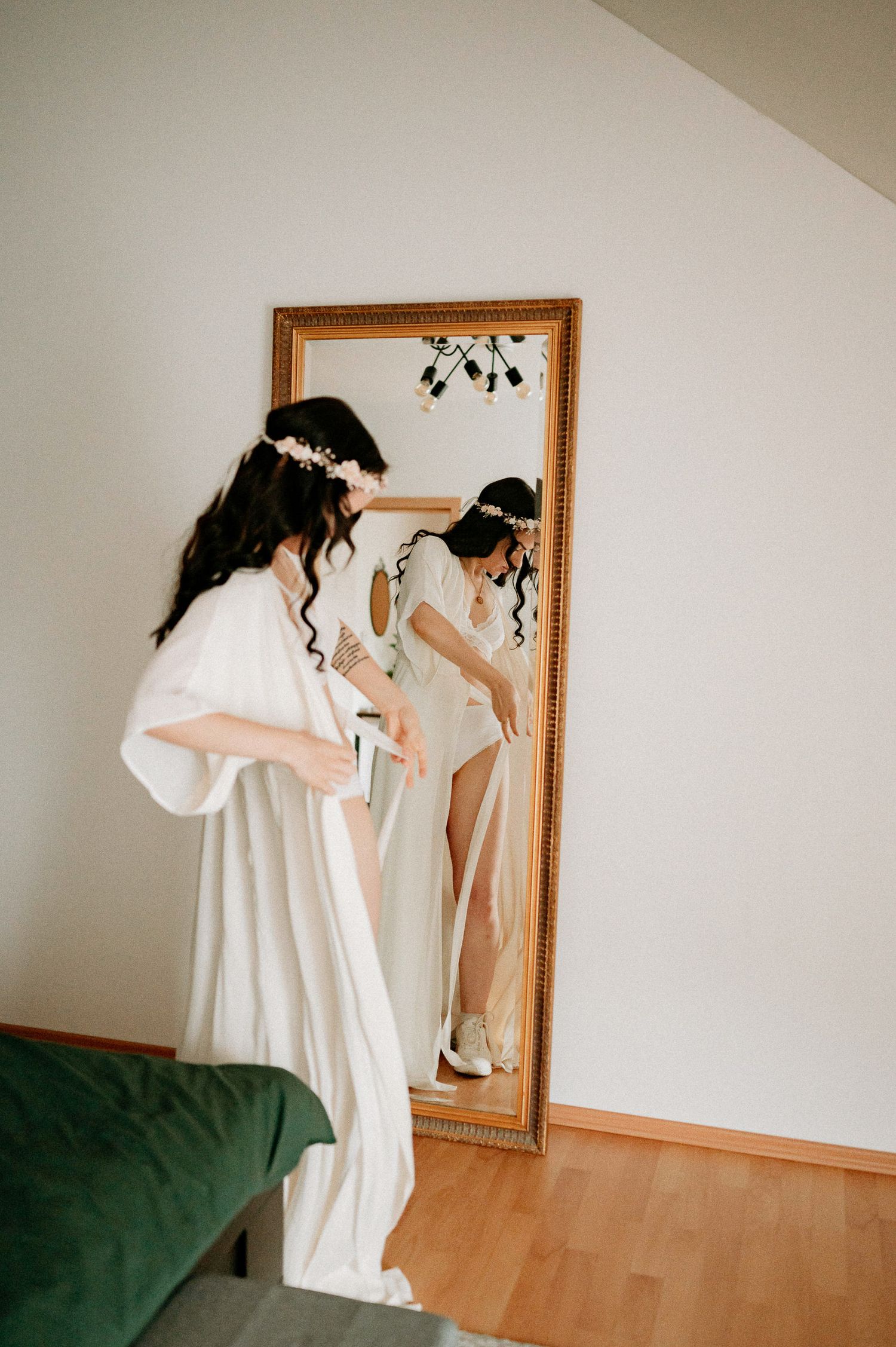 A figure in a flowing white dress poses contemplatively in front of a gold-framed mirror in a minimalist room.