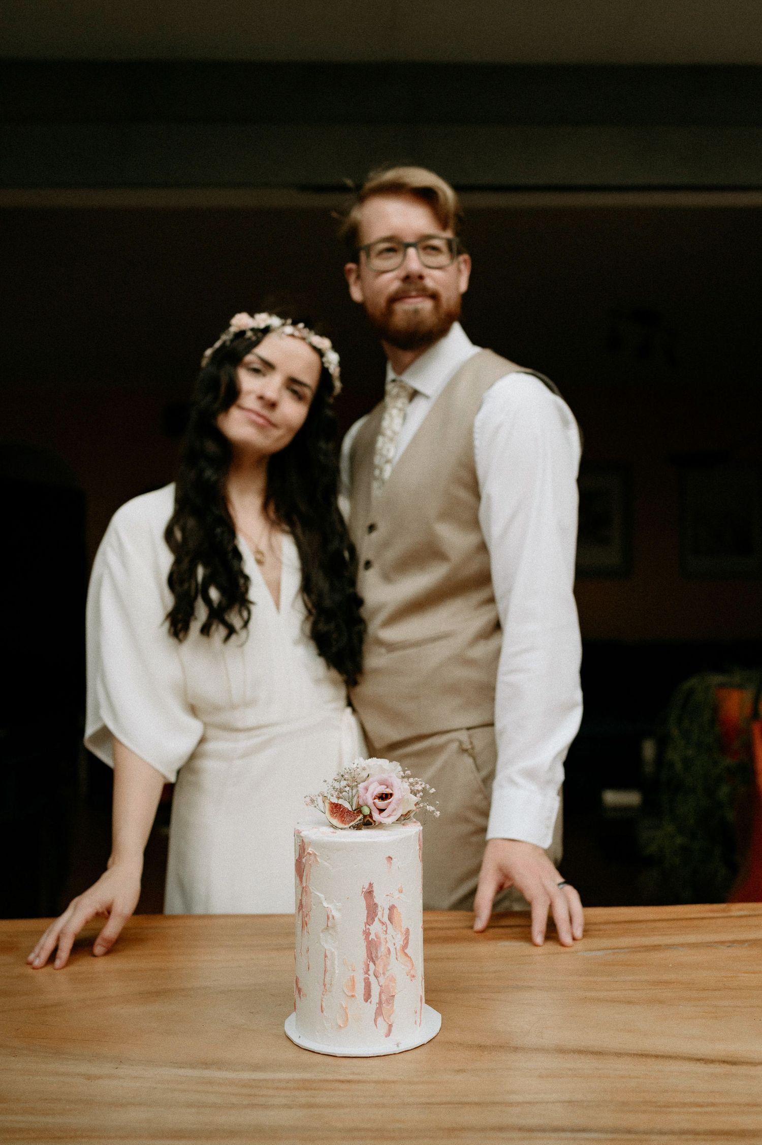 Couple poses casually at wooden table with their decorated wedding cake.