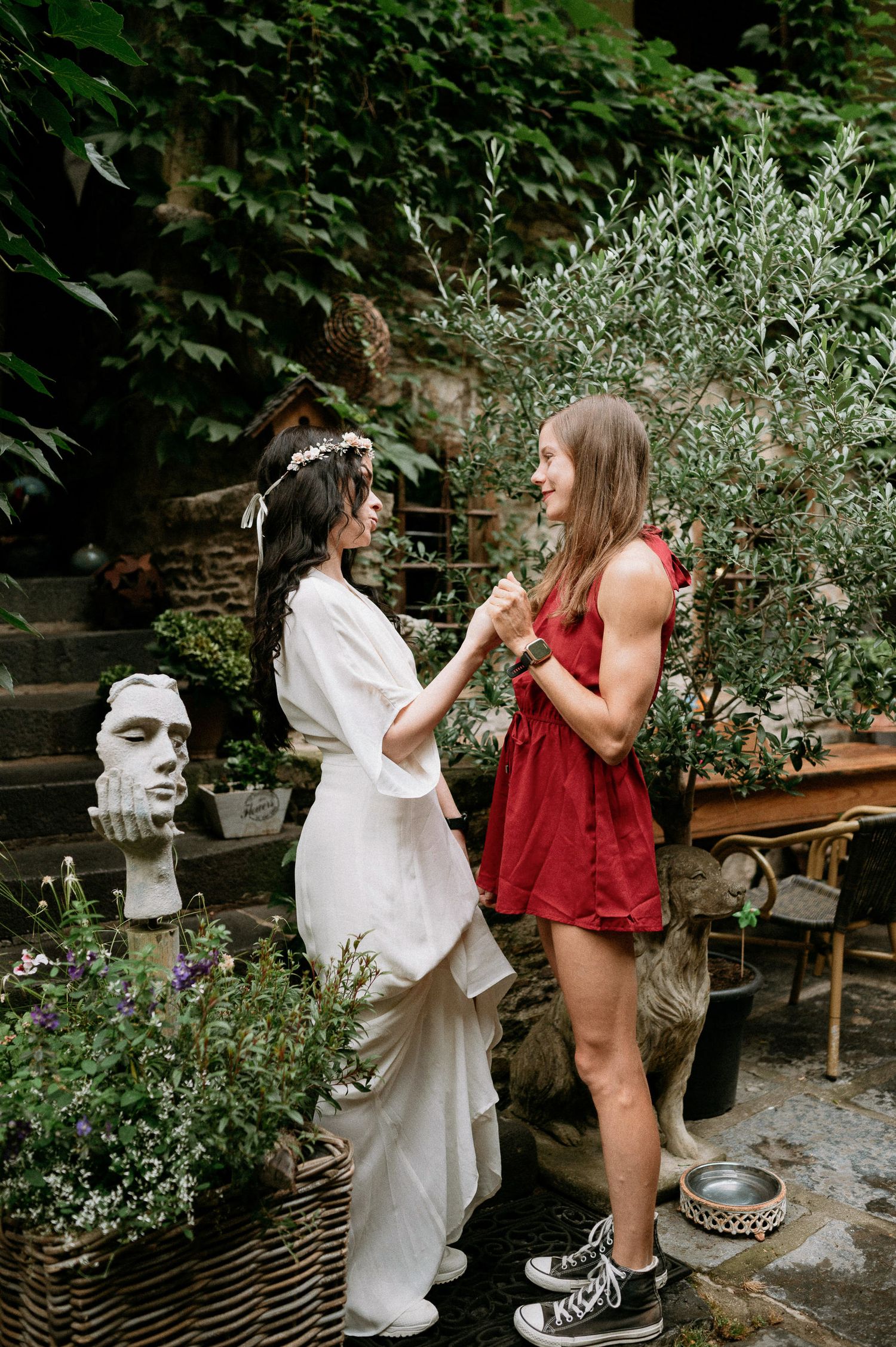 Two people share a moment in a garden with decorative stone sculptures and greenery.