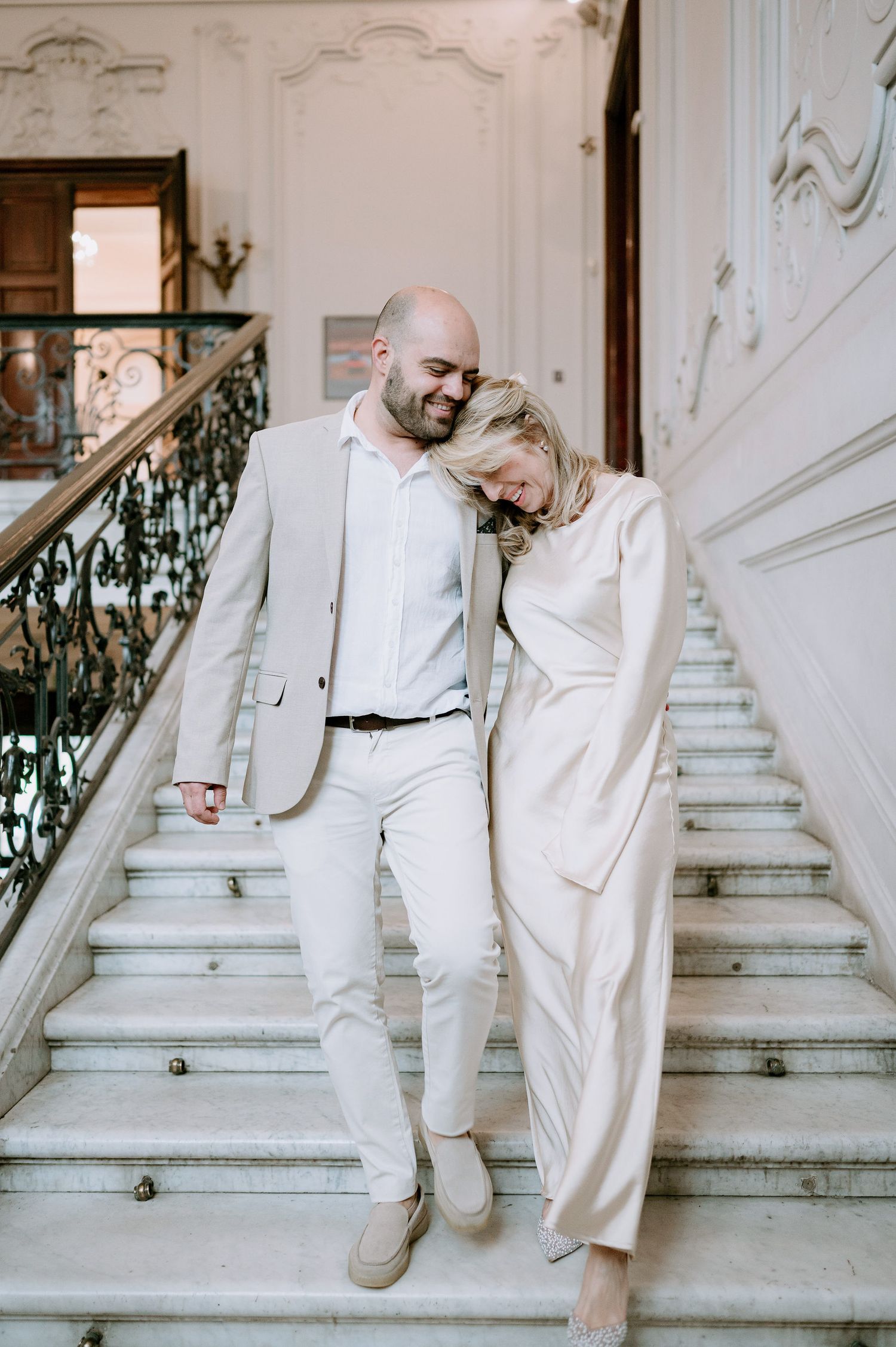 A couple in matching light-colored outfits embrace on marble stairs with decorative iron railings.