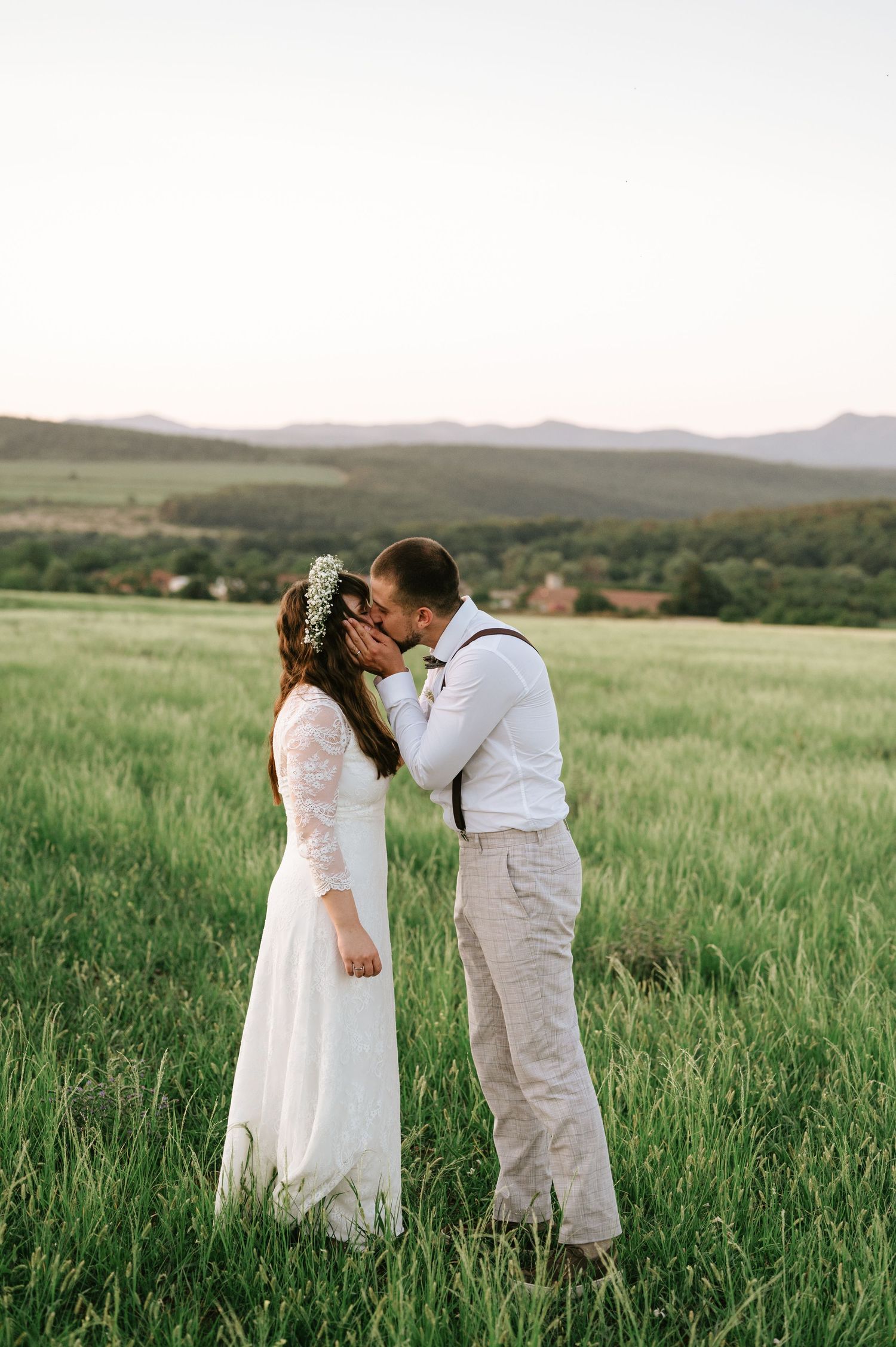 Newlyweds share intimate moments in a vast green meadow with mountains in the background.
