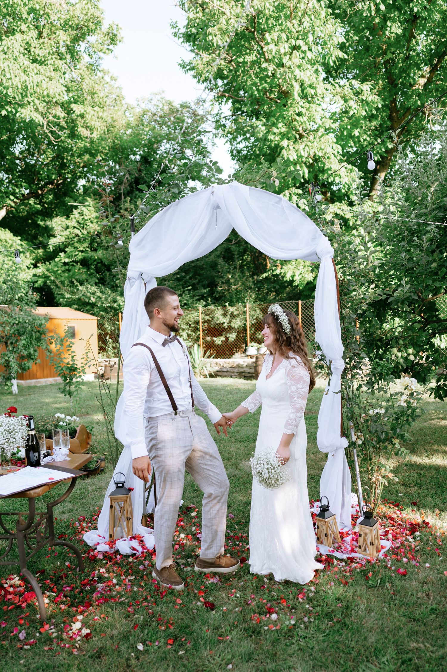 Couple sharing a joyful moment under an elegant white wedding arch decorated with flowers in a garden setting.