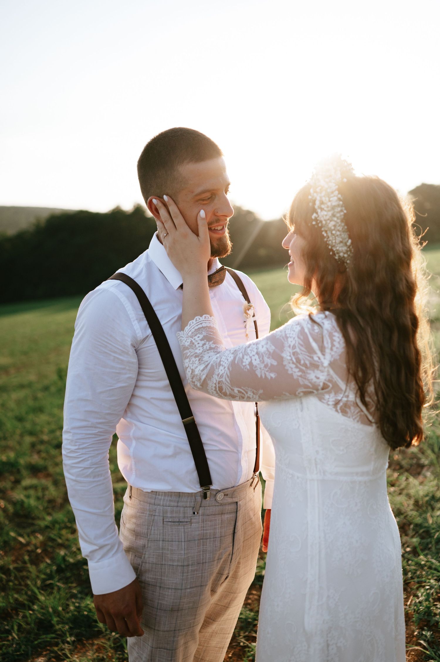 Bride in lace gown caresses groom's face during sunset wedding photoshoot.