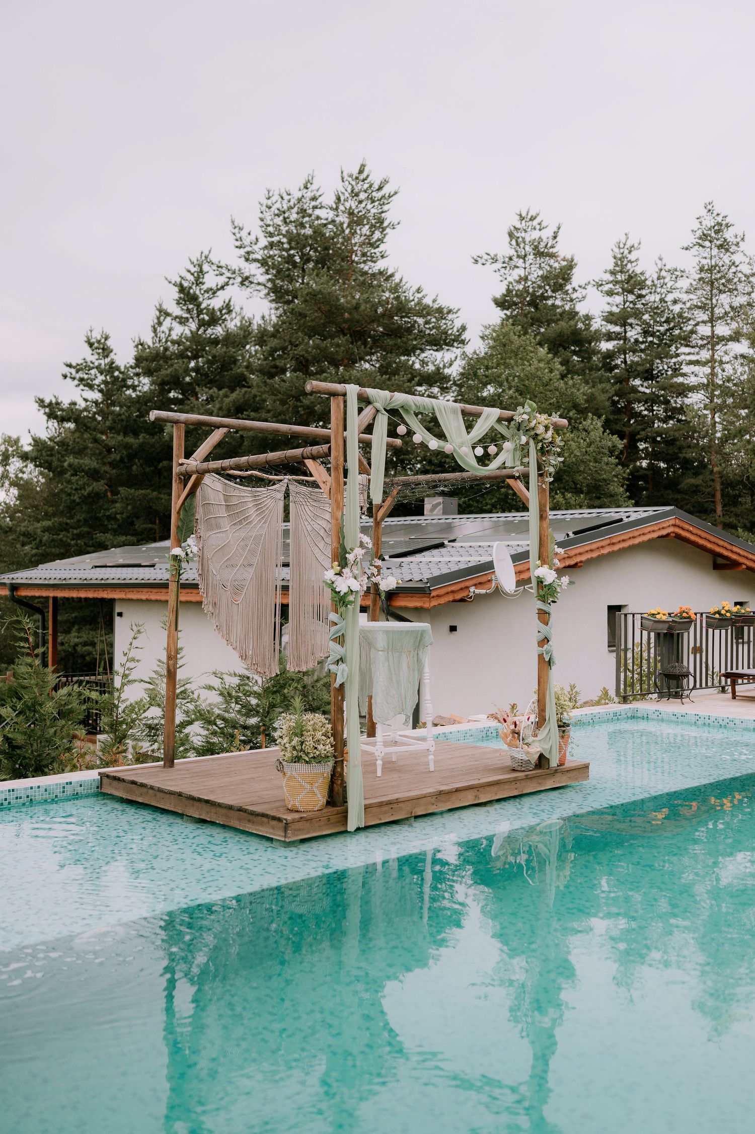A modern pool deck features a wooden pergola and turquoise water surrounded by tall pine trees.