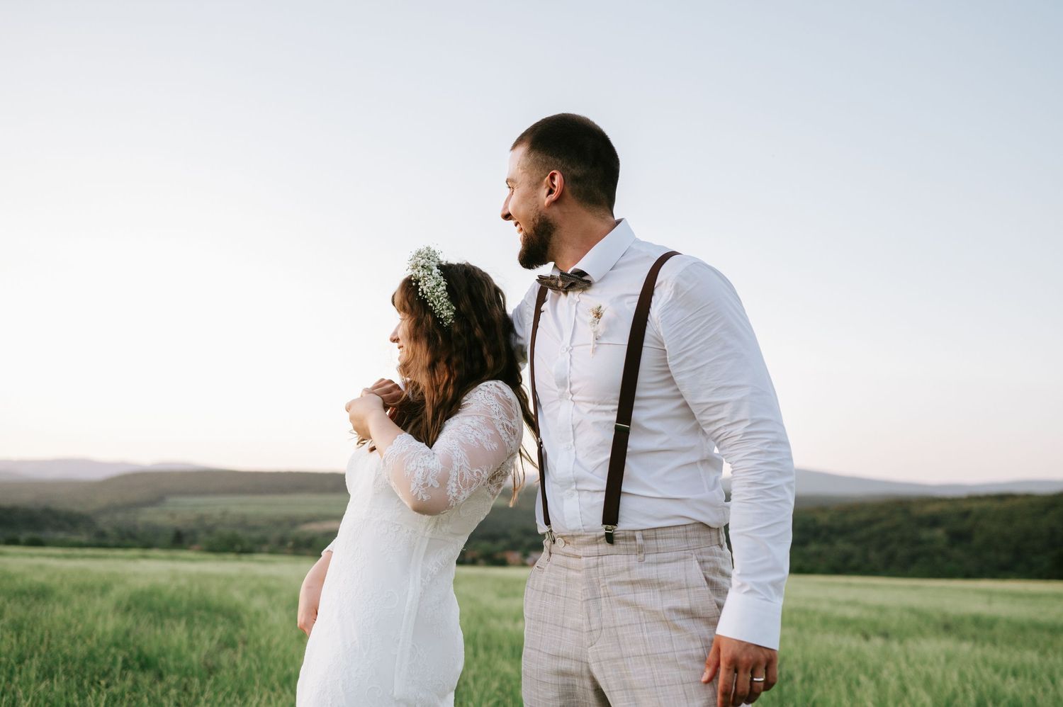 Couple in white and suspenders walking through a scenic meadow at sunset.