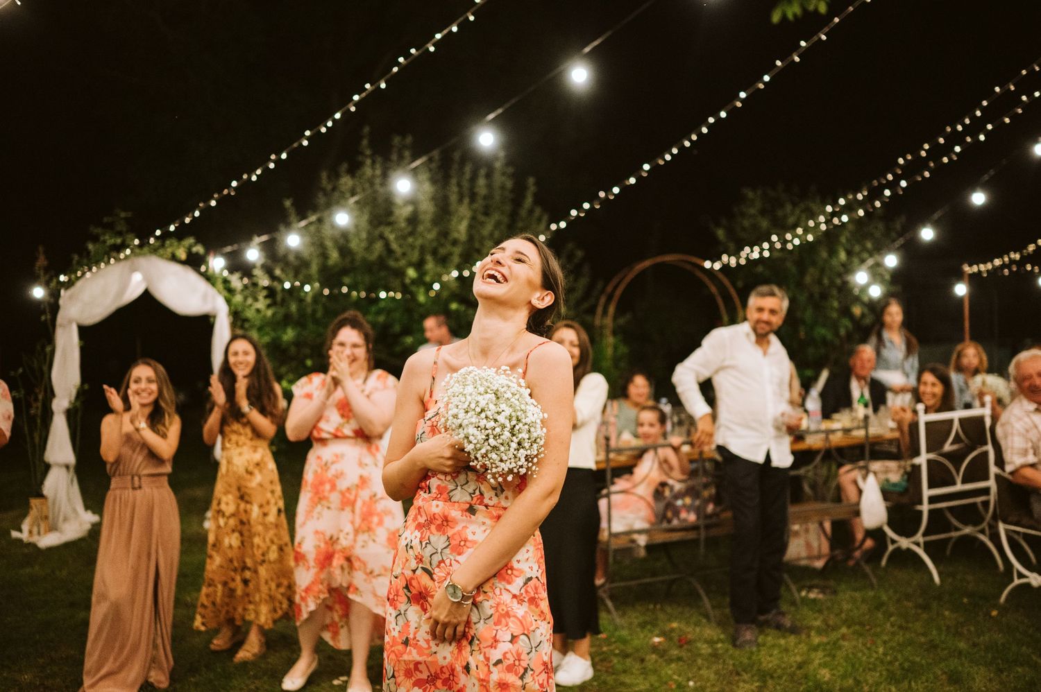 String lights illuminate an outdoor wedding reception as guests celebrate on the lawn.