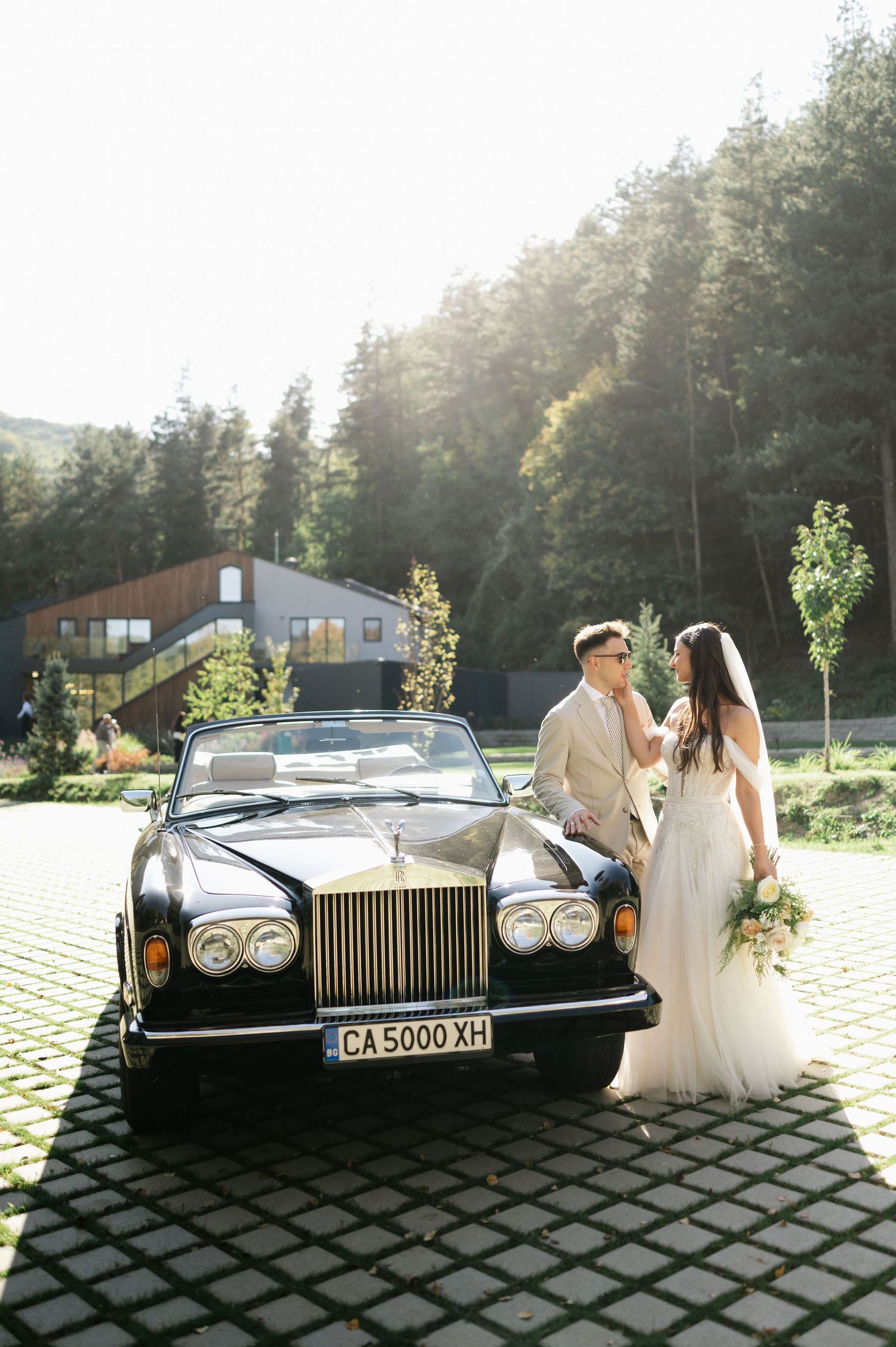 Newlyweds share a tender moment beside a luxury vintage car with mountains and forest in the background.