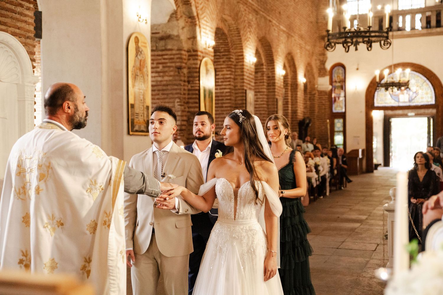Orthodox wedding ceremony in a historic brick church with arched columns and ornate chandeliers.