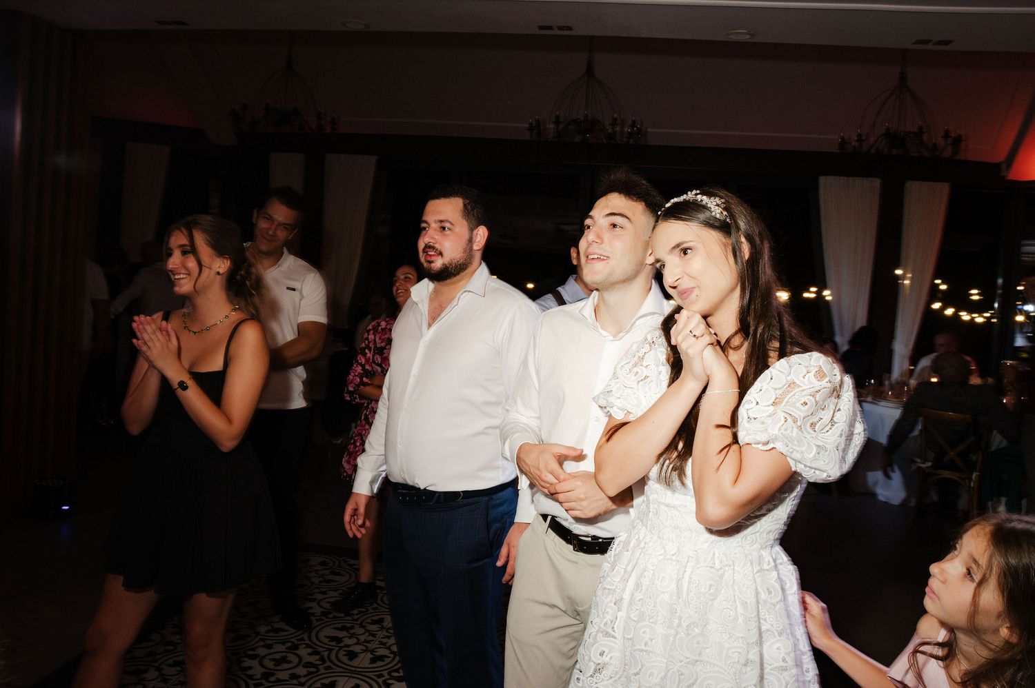 Party guests react excitedly while watching an event at an indoor reception venue with dim lighting.