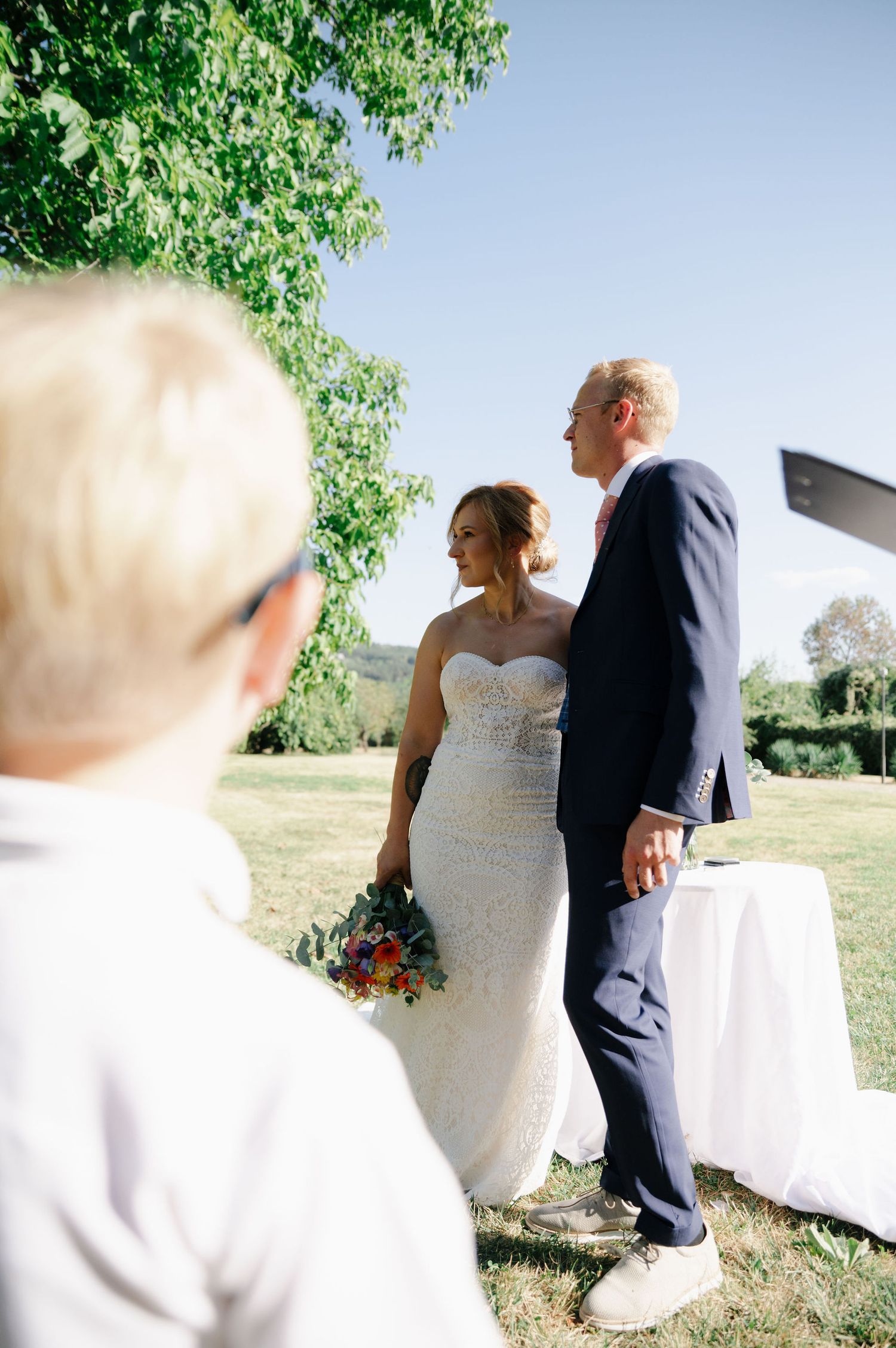 Wedding couple stands together in an outdoor setting with trees in background.