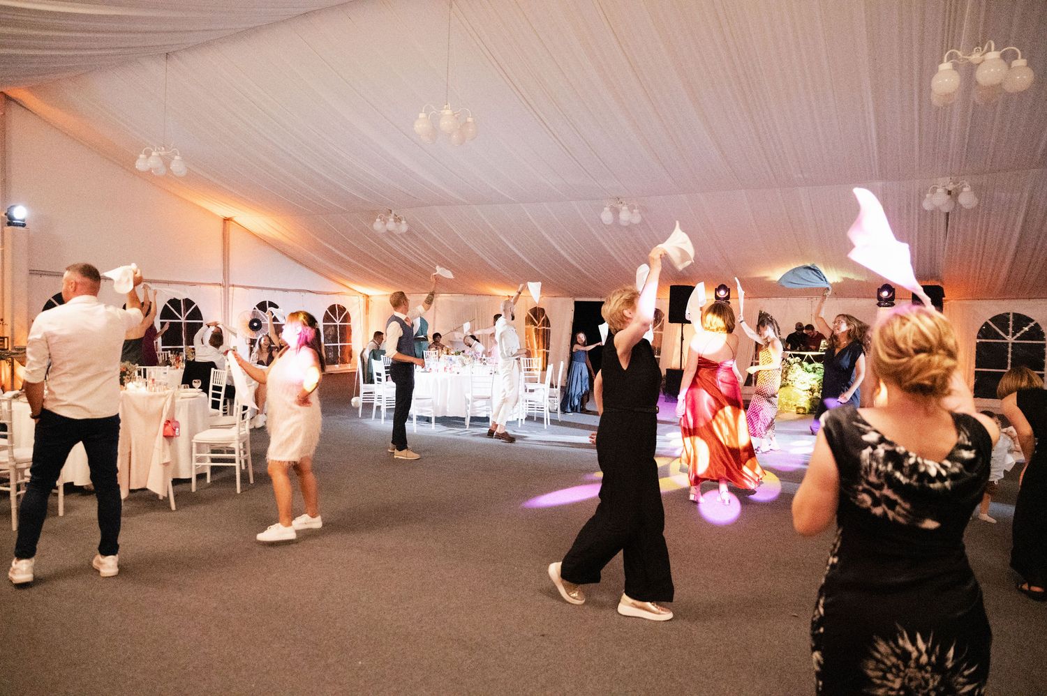 People dancing and celebrating at an elegant wedding reception in a white tent with glowing lanterns and ambient lighting.