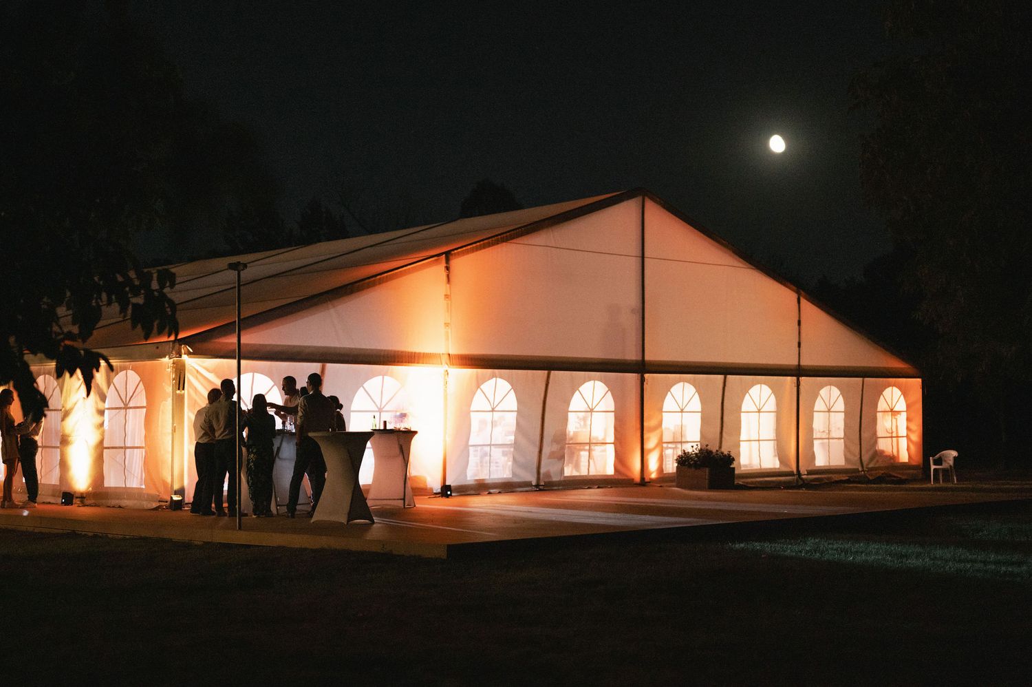 An illuminated white party tent glows warmly at night with silhouettes of guests visible through arched windows.
