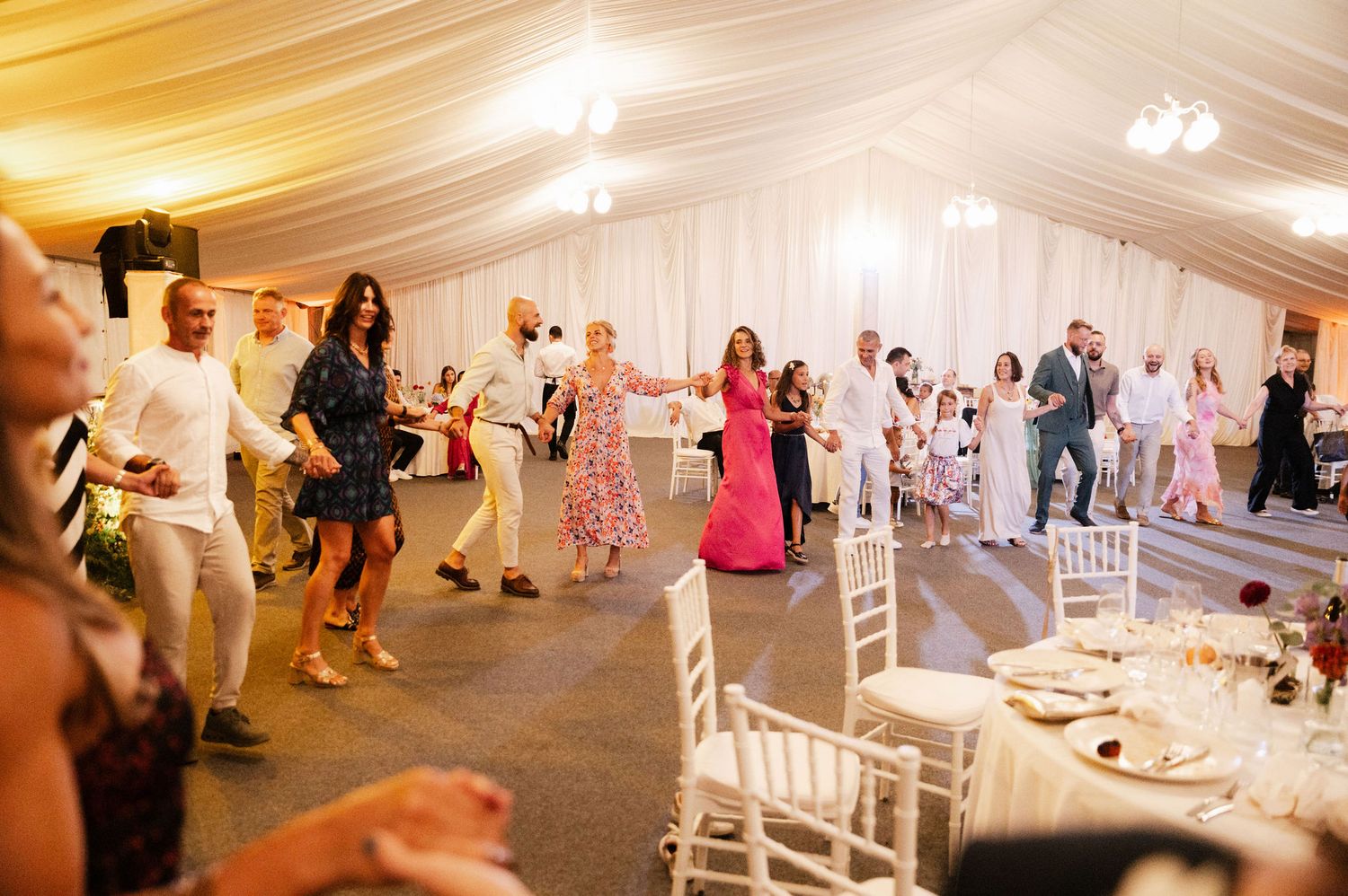 Wedding guests dance and celebrate together on an illuminated dance floor inside a white tent reception venue.