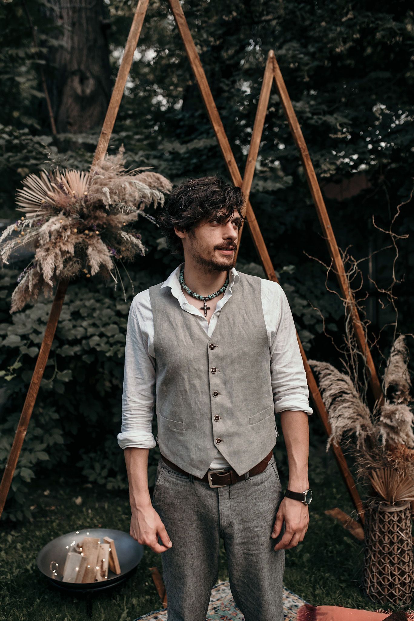 Person wearing vintage-style gray vest and white shirt standing near wooden structures and dried flowers.