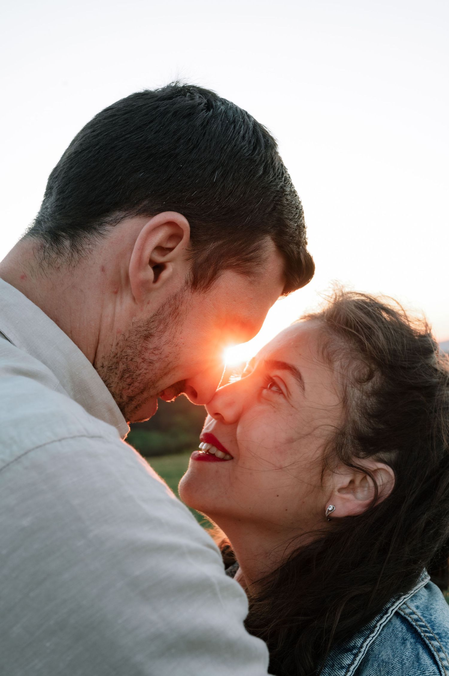 A romantic couple shares an intimate moment at sunset with sunlight flaring between them as they smile at each other.