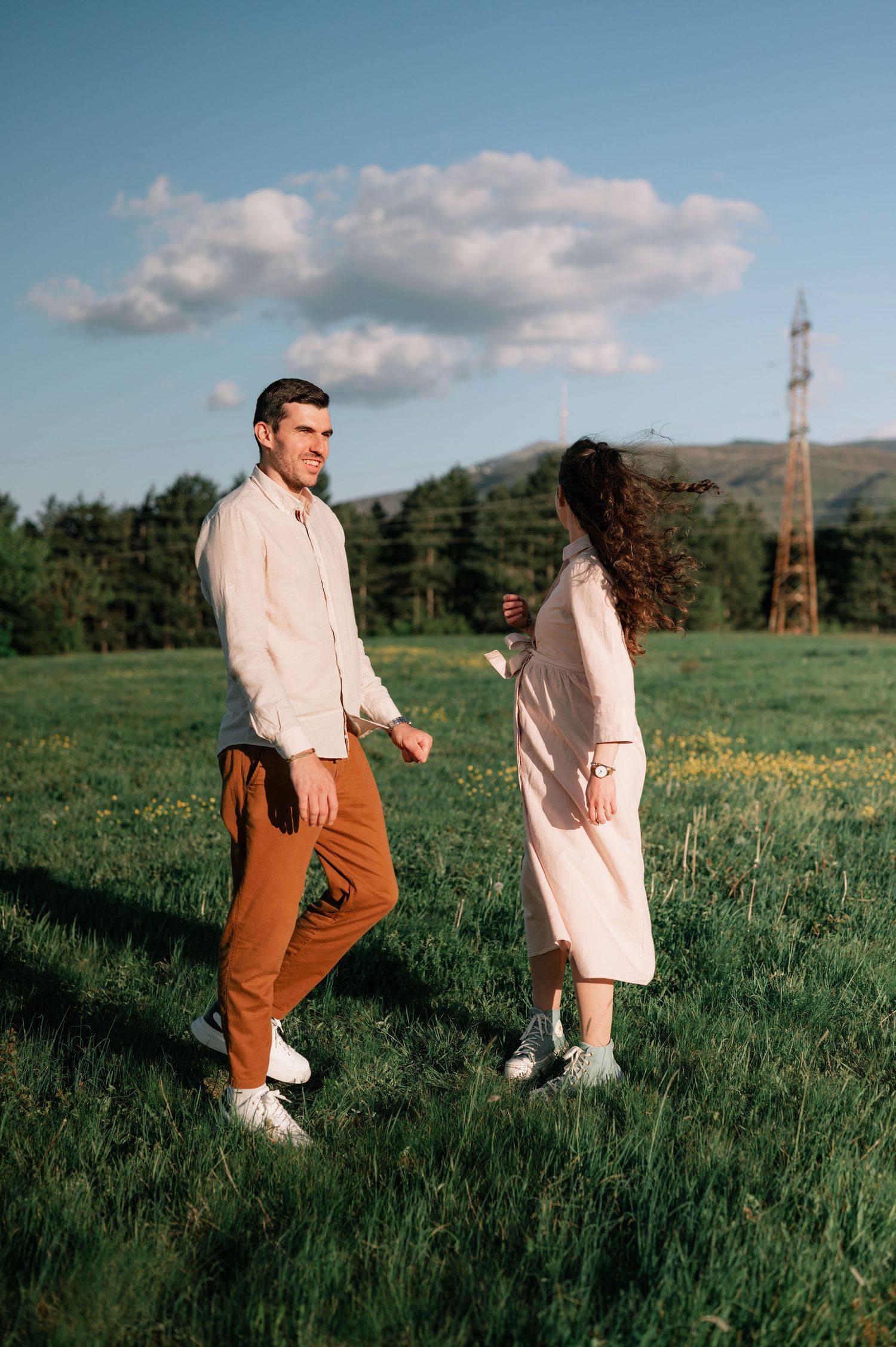 Two people in casual attire walk through a sunny green meadow with mountains and blue sky in the background.