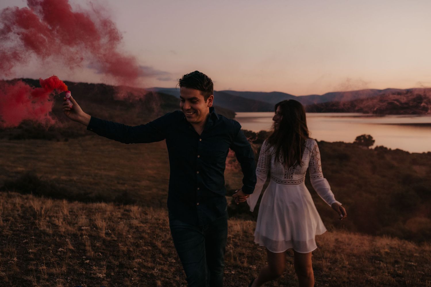 Romantic couple poses on a hillside at dusk with red smoke creating an ethereal atmosphere.