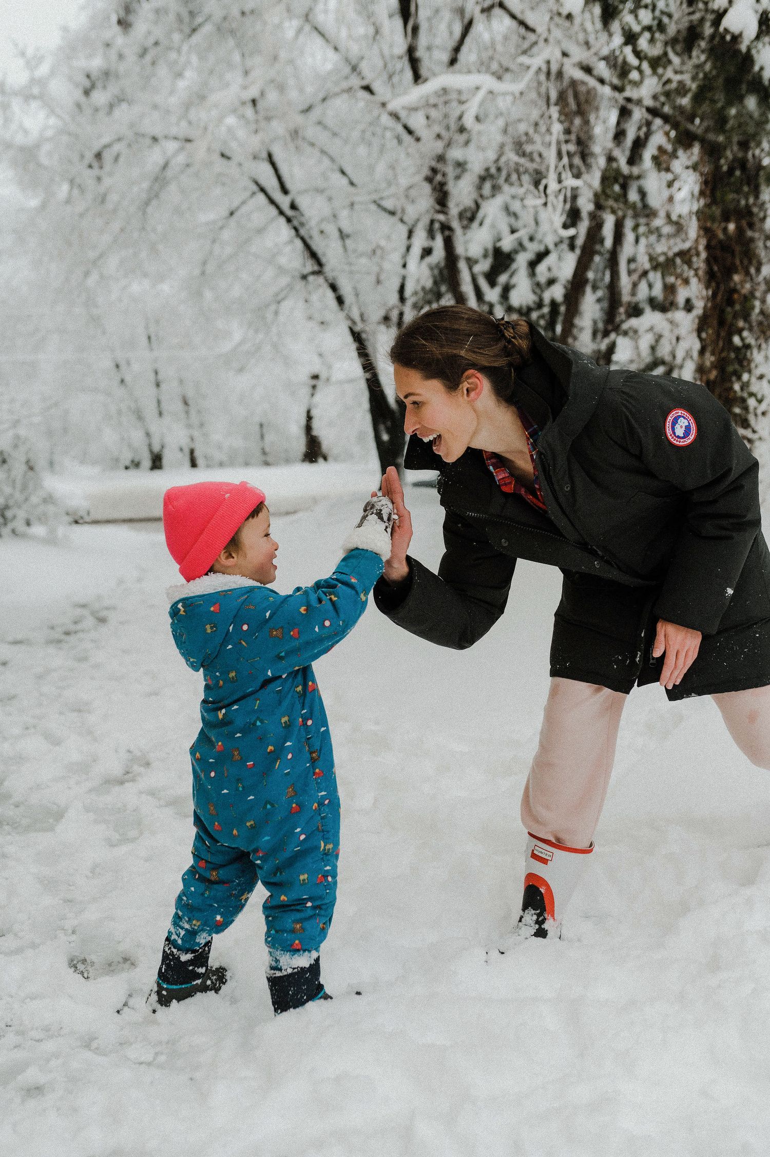 A small child in a blue snowsuit plays in deep winter snow while an adult in black winter clothing watches nearby.