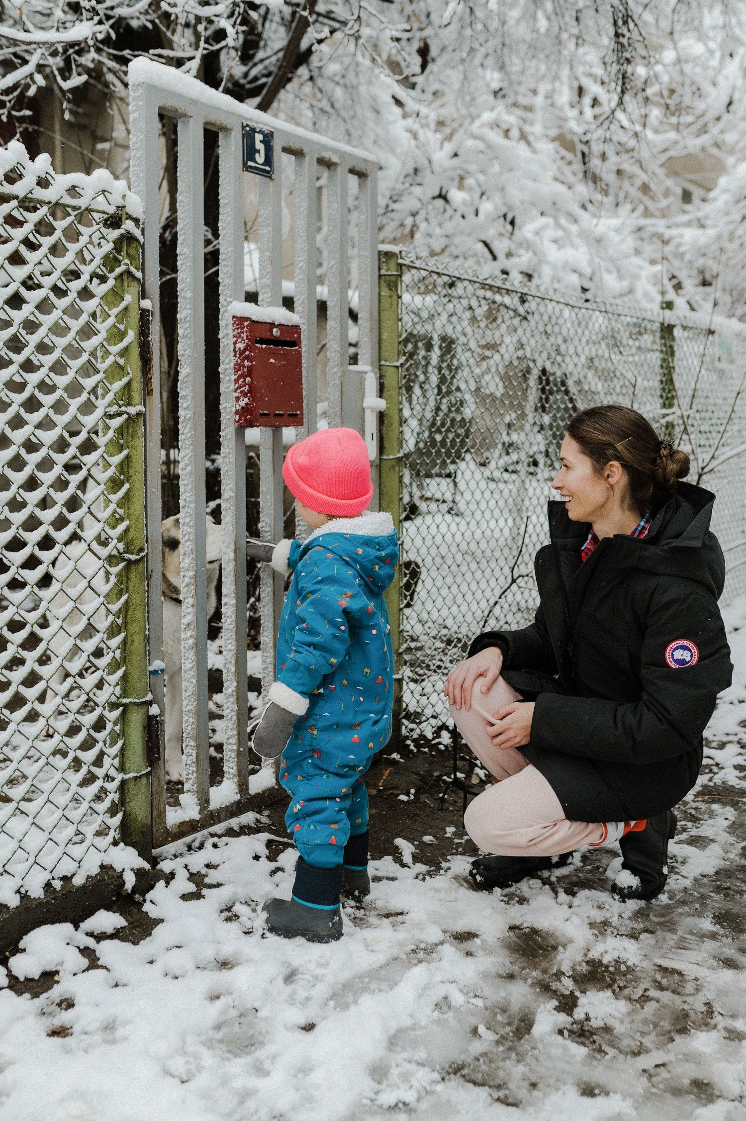 A child in a blue snowsuit explores a snowy fence while someone in a black winter coat watches nearby.