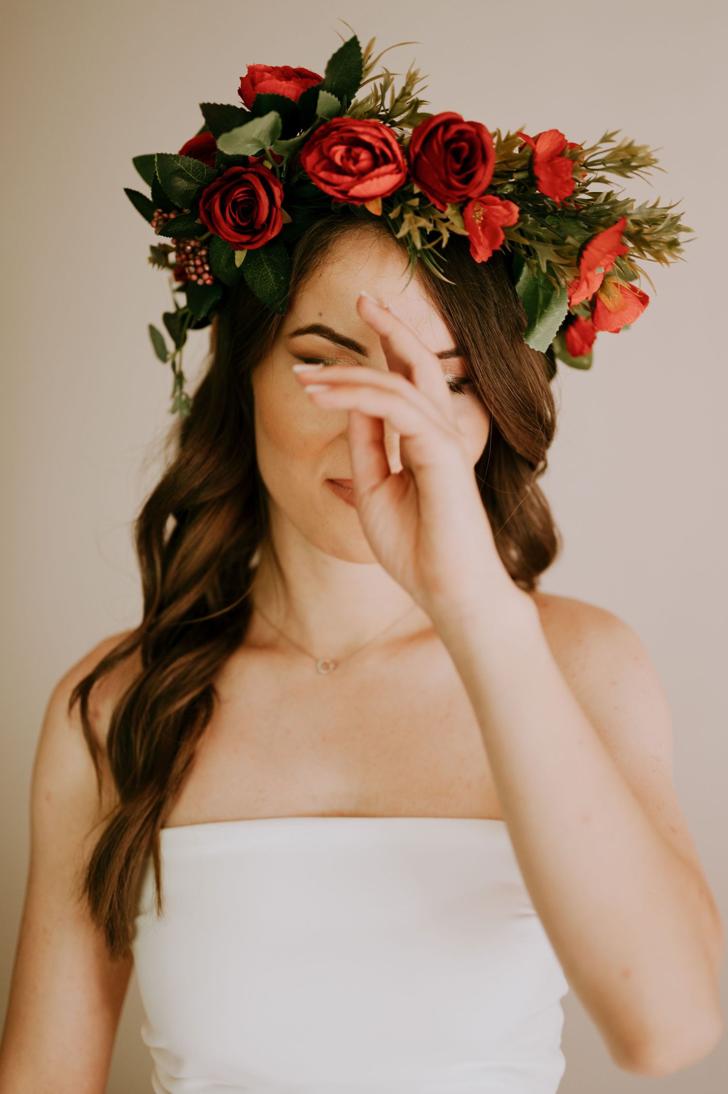 Playful bridal portrait showing white dress and red rose crown with hands covering face.