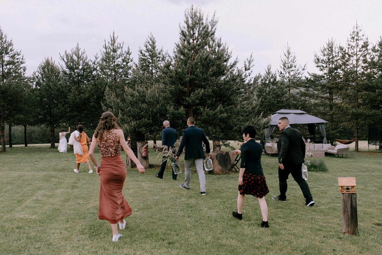 People dancing and celebrating outdoors on a grassy field surrounded by pine trees.