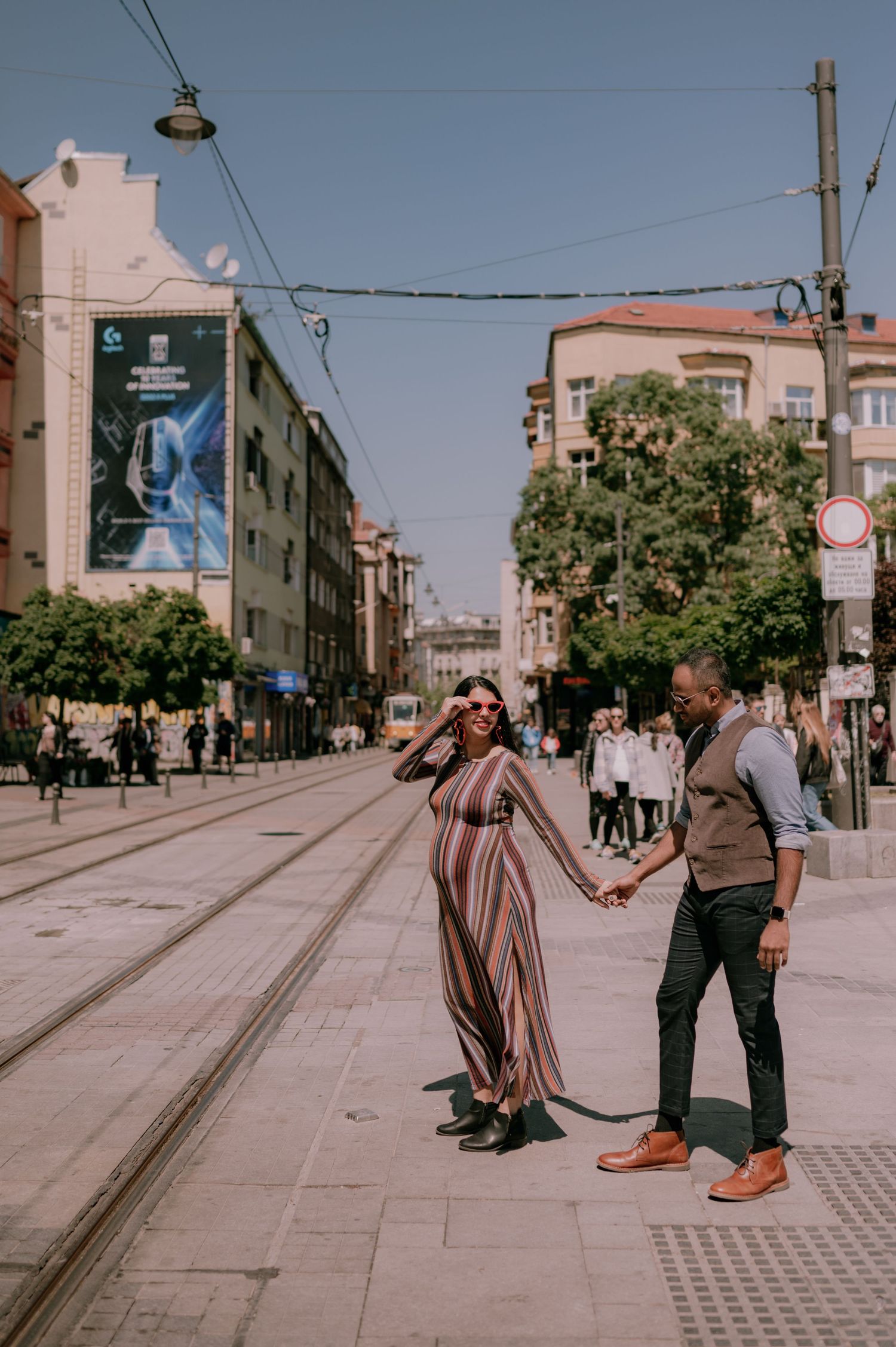 A couple holds hands and walks along a tram track on a sunny urban street, woman in striped maxi dress and sunglasses.
