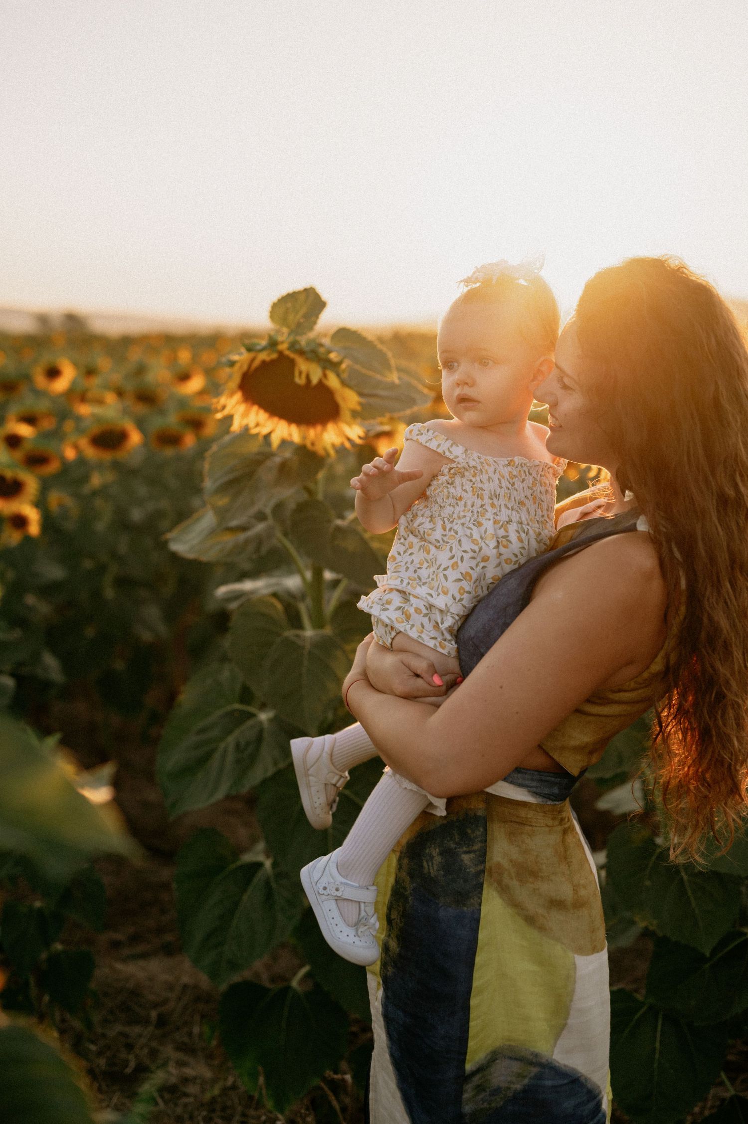 Mother holding baby girl in sunflower field at golden hour sunset, warm light glowing behind them.