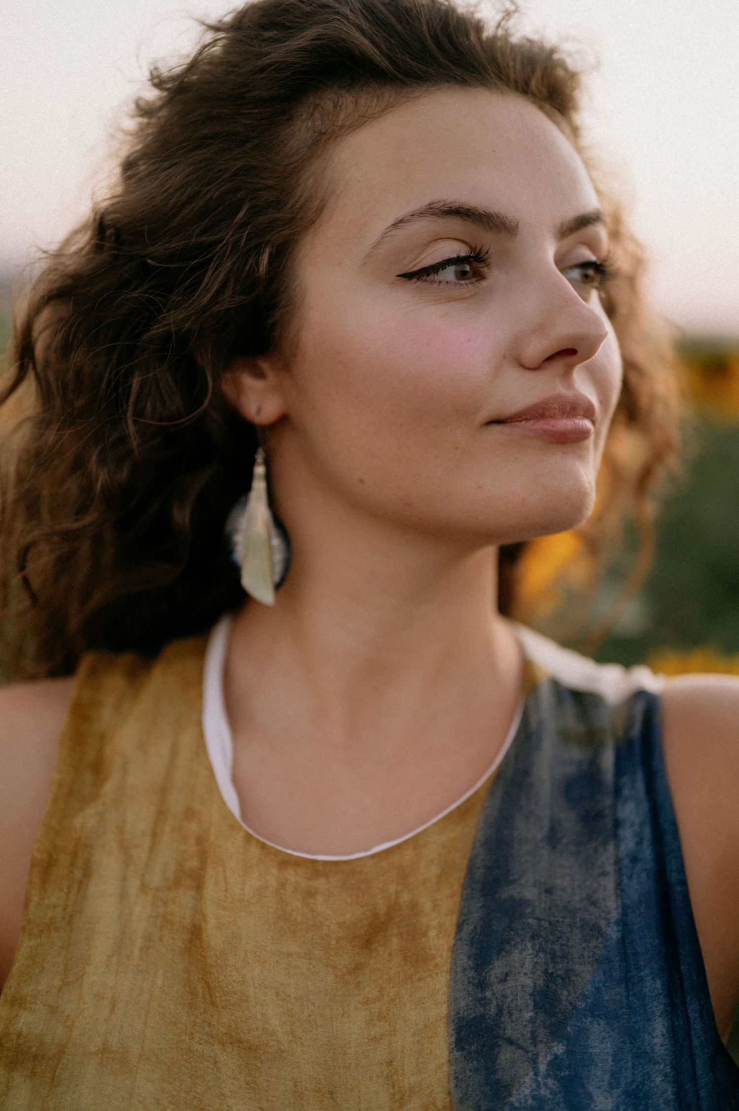 Young woman with curly hair wearing feather earrings and a gold and blue top, bathed in warm golden hour sunlight.
