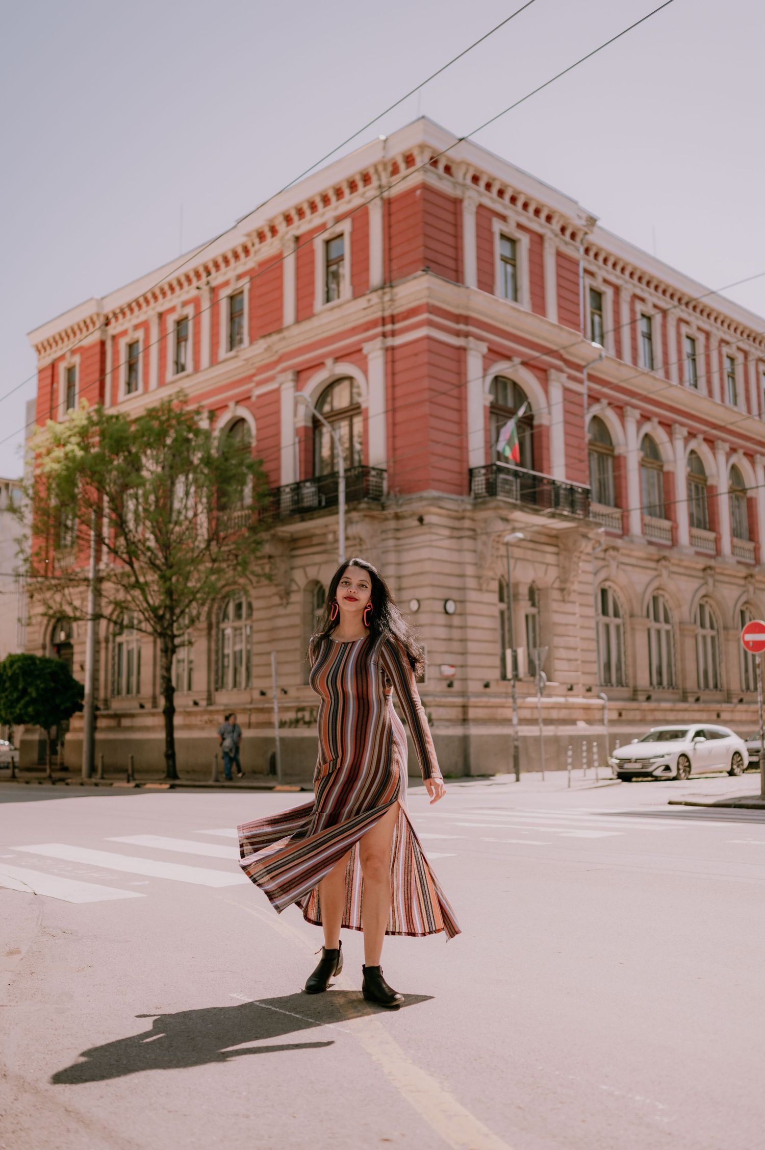 Woman in striped midi dress posing on sunny street corner near elegant red historic building.