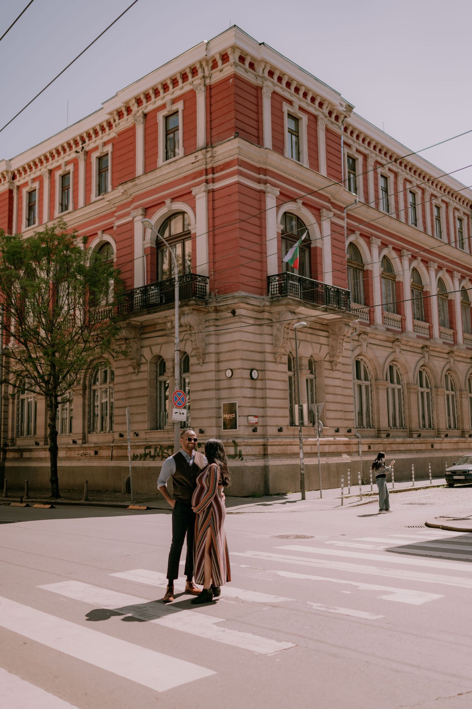 Couple embracing on a crosswalk in front of a grand red neoclassical building on a sunny urban street.