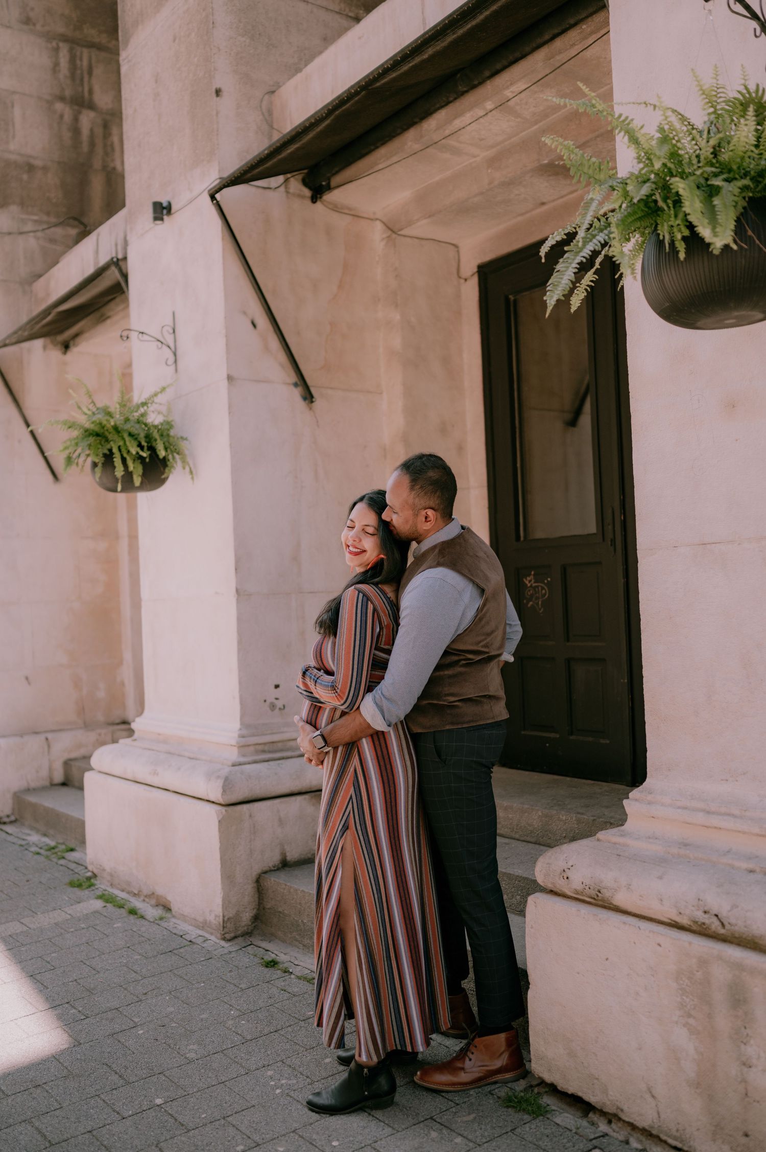 Couple embracing outside a historic building with hanging ferns, woman in striped dress smiling as man kisses her cheek.