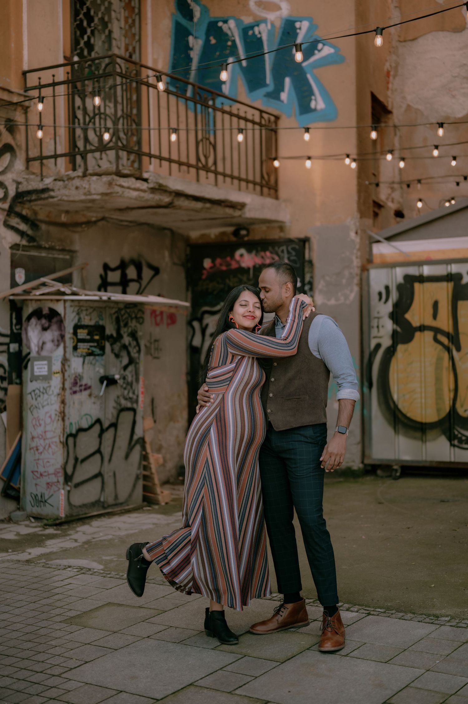 Couple embracing romantically on a urban street with graffiti walls and string lights in the background.
