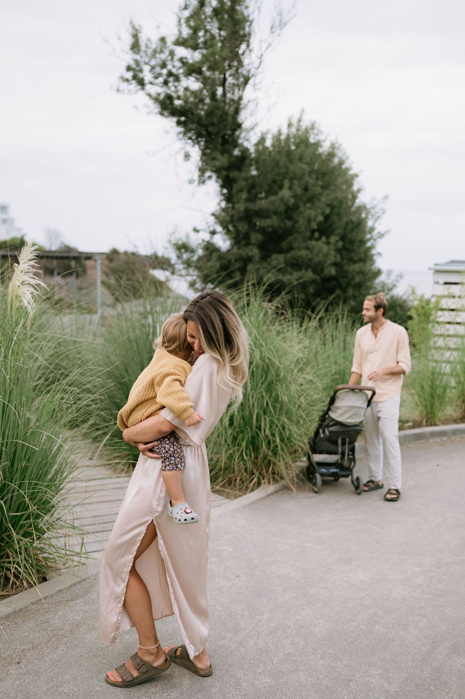 A woman in a pink dress holds a toddler outdoors while a man pushes a stroller on a paved path.