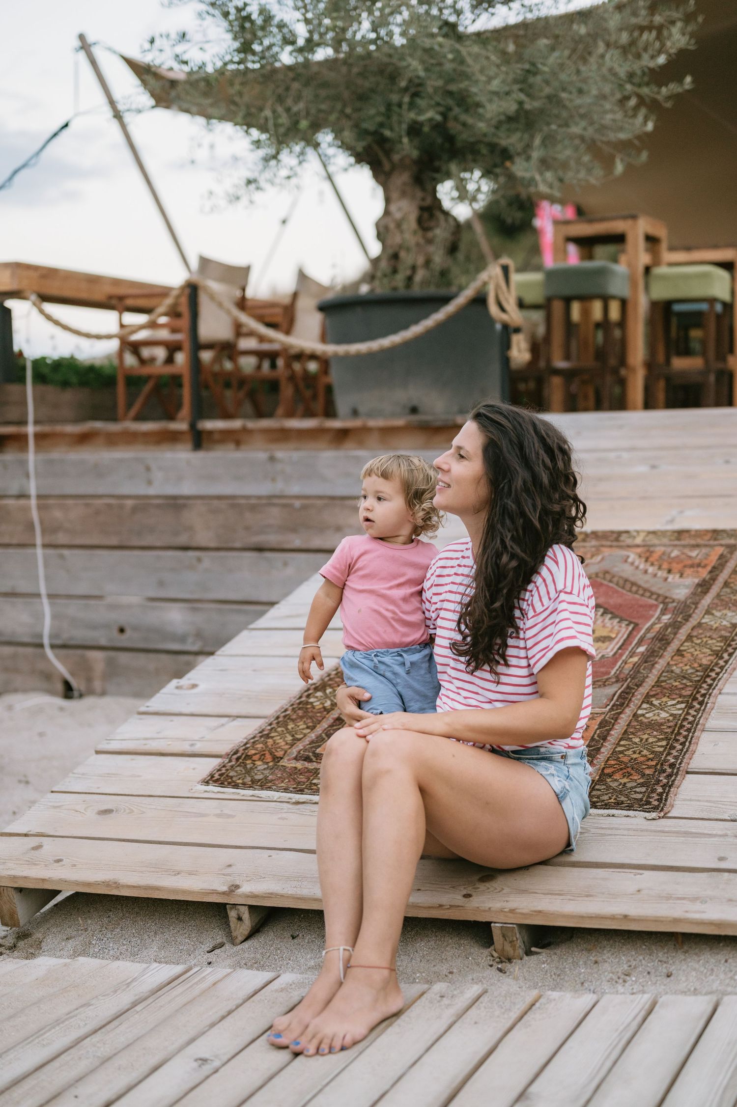 A young mother with dark curly hair sits on a wooden deck holding her toddler, both wearing pink striped shirts.