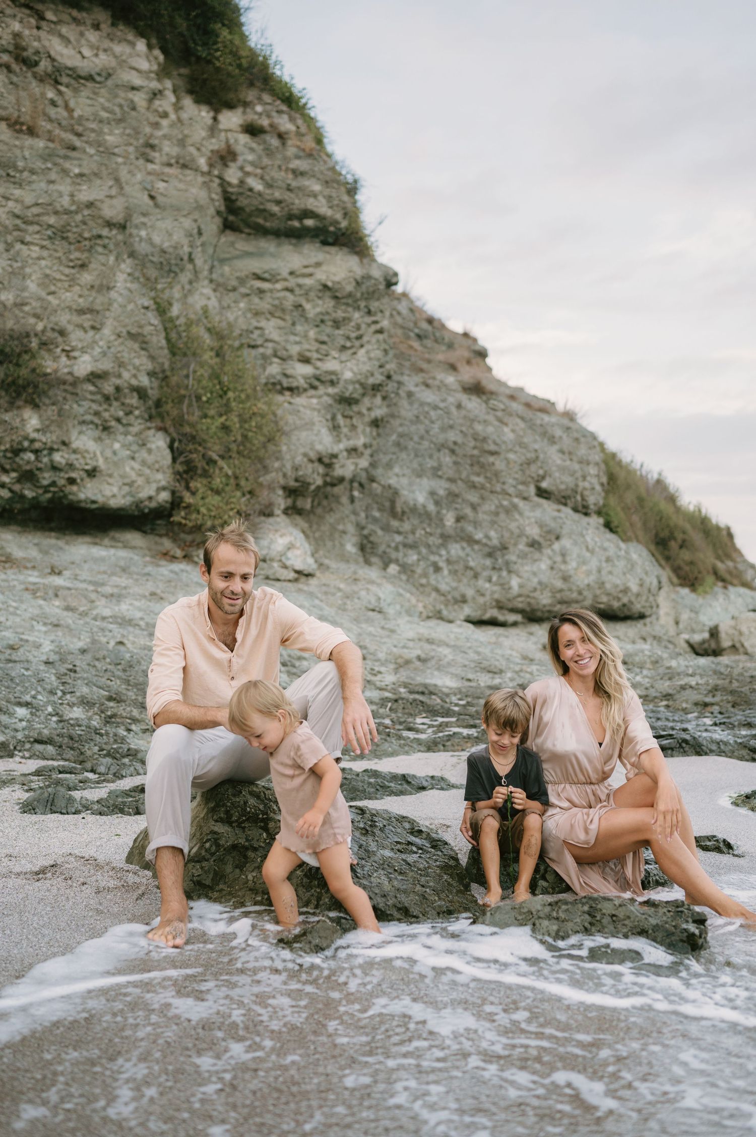 Family of four sitting on rocks at a sandy beach with a rocky cliff backdrop, enjoying a relaxed coastal outing.