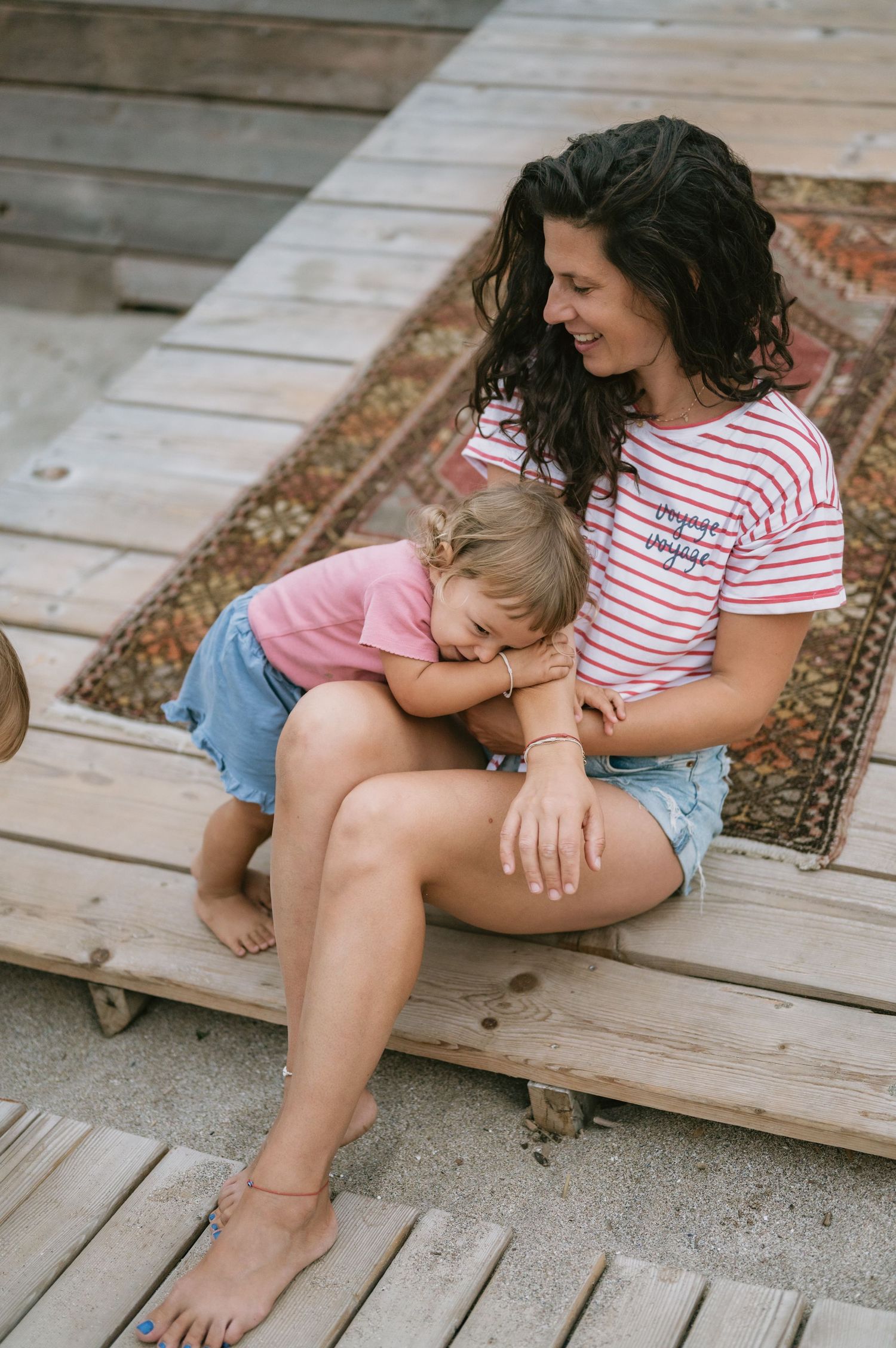 A smiling mother in a striped shirt holds her young daughter on wooden steps at a beach.